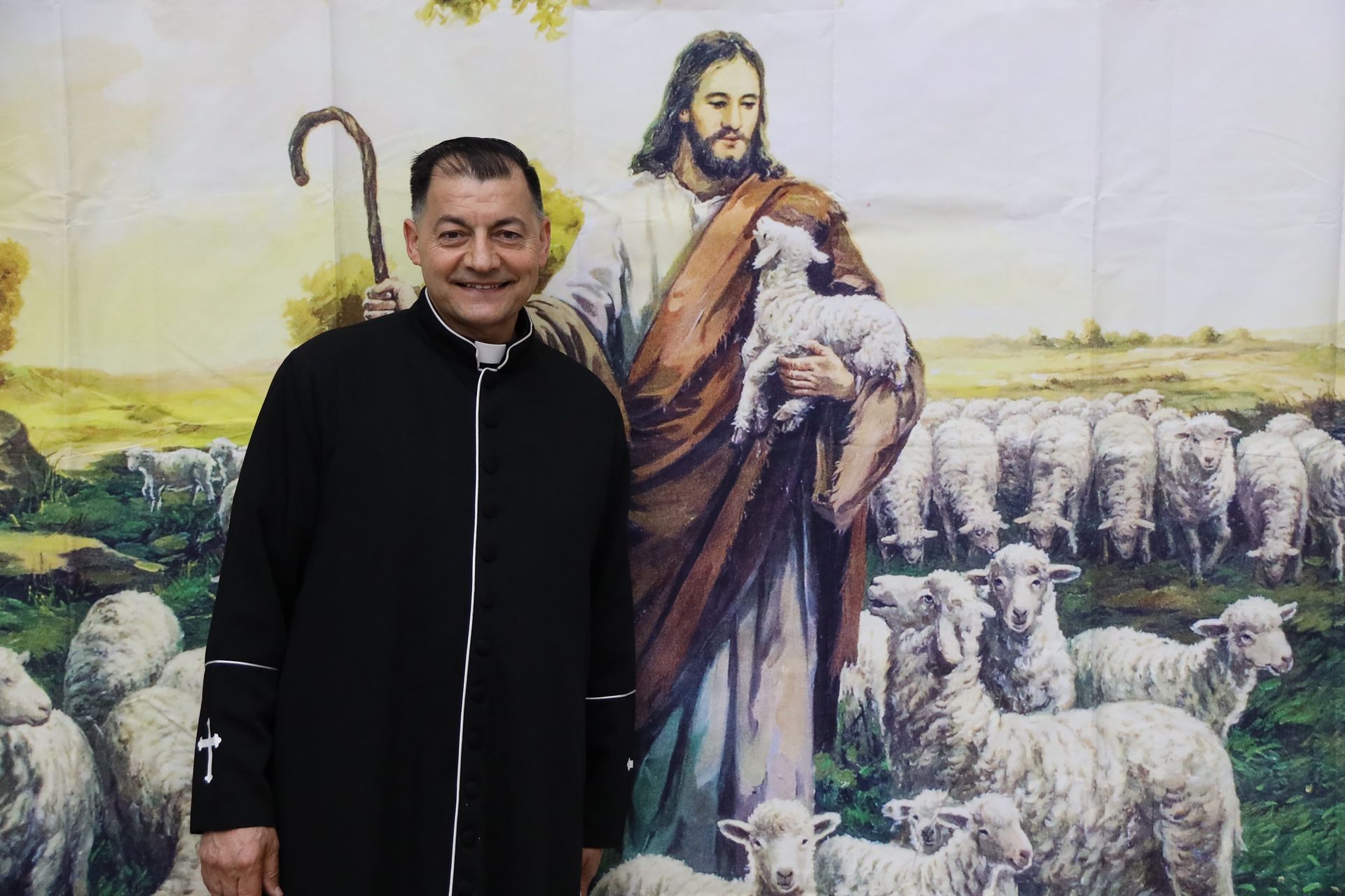 Smiling man in a black coat stands before a pastoral mural of Jesus with sheep