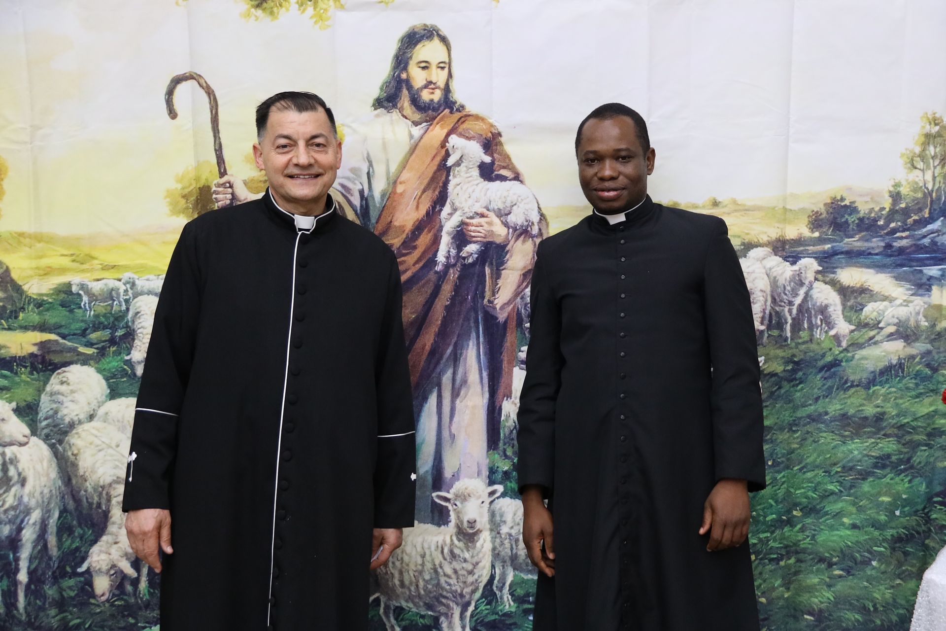 Two men in black robes standing before a mural of Jesus in a pastoral landscape