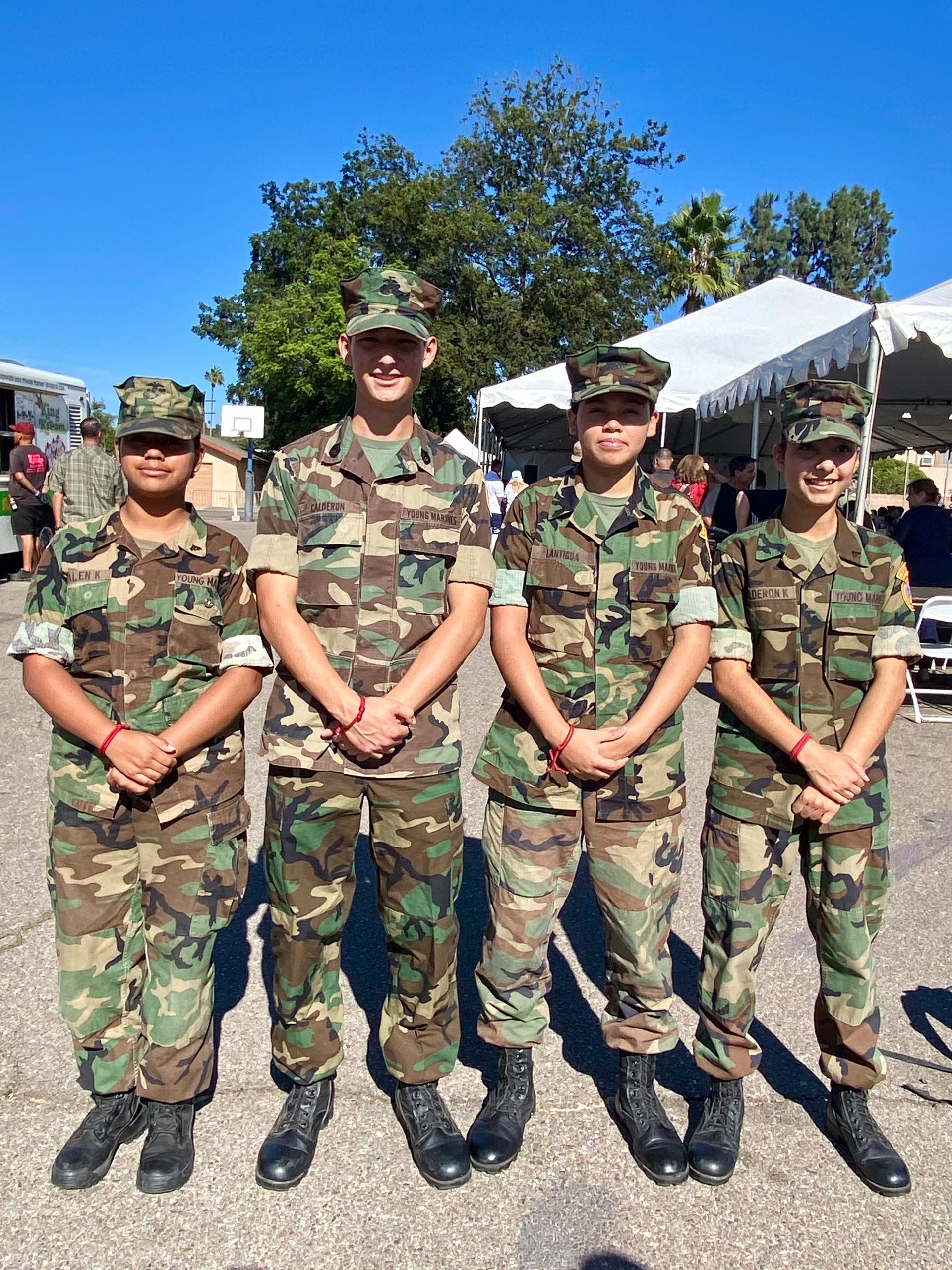 Four people in camouflage uniforms stand smiling outdoors in front of tents on a sunny day.
