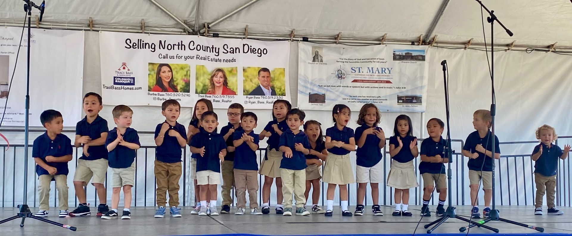 Children in matching blue shirts and white shorts performing on a stage under a tent