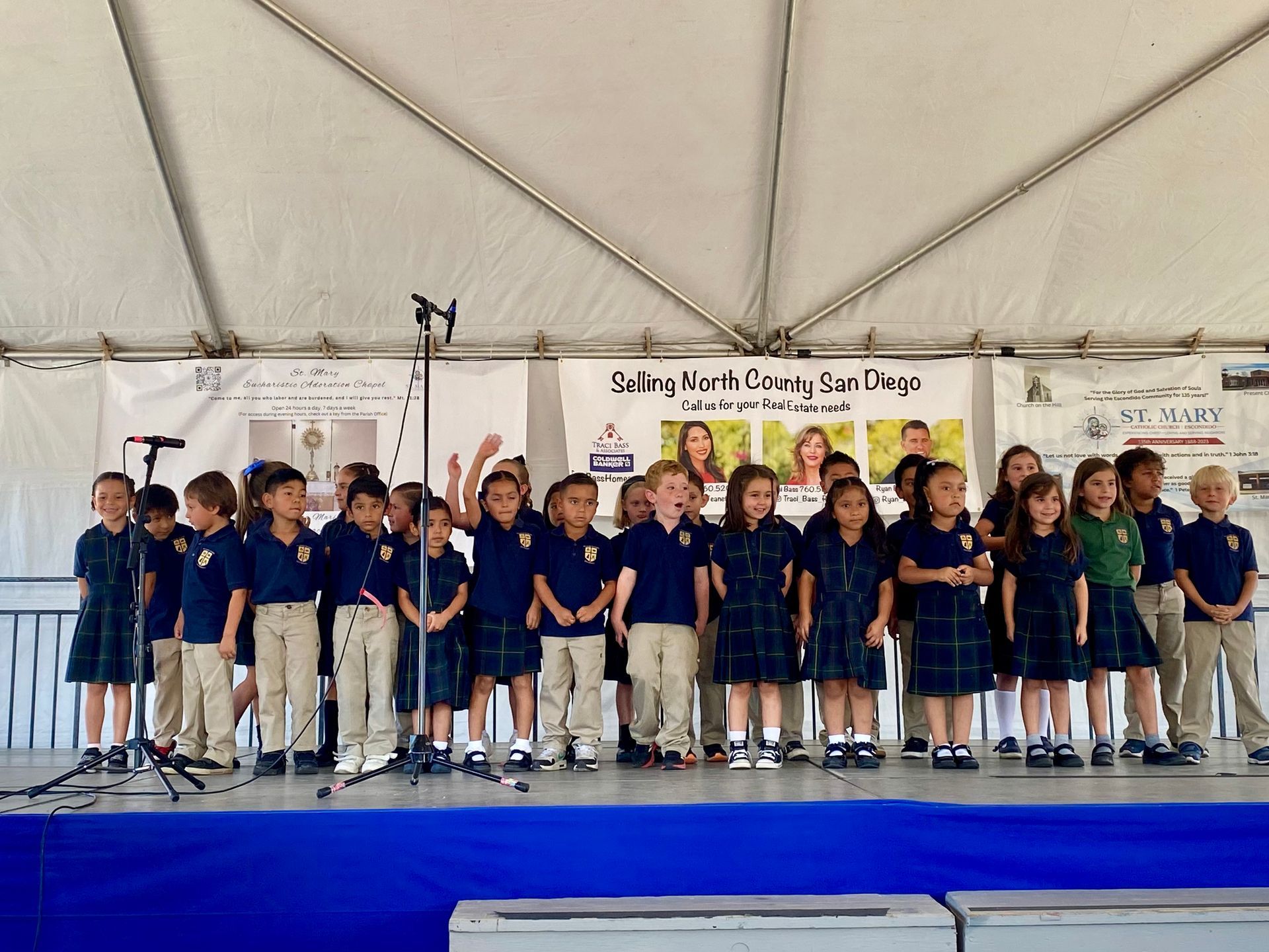 Group of children in navy shirts standing on a stage under a tent, with banners behind them