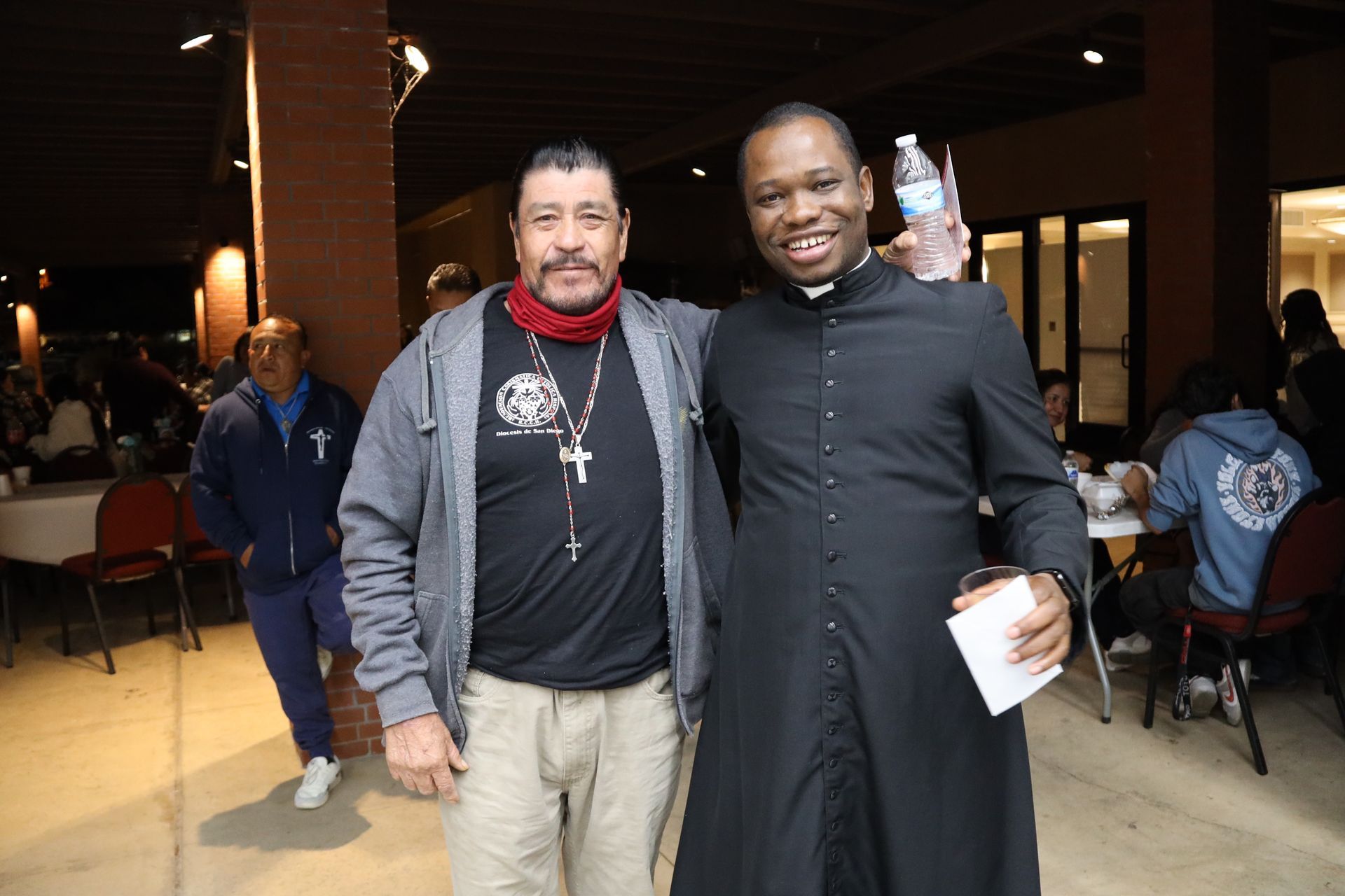 Two men standing together indoors, one holding a water bottle and paper, in a banquet hall.