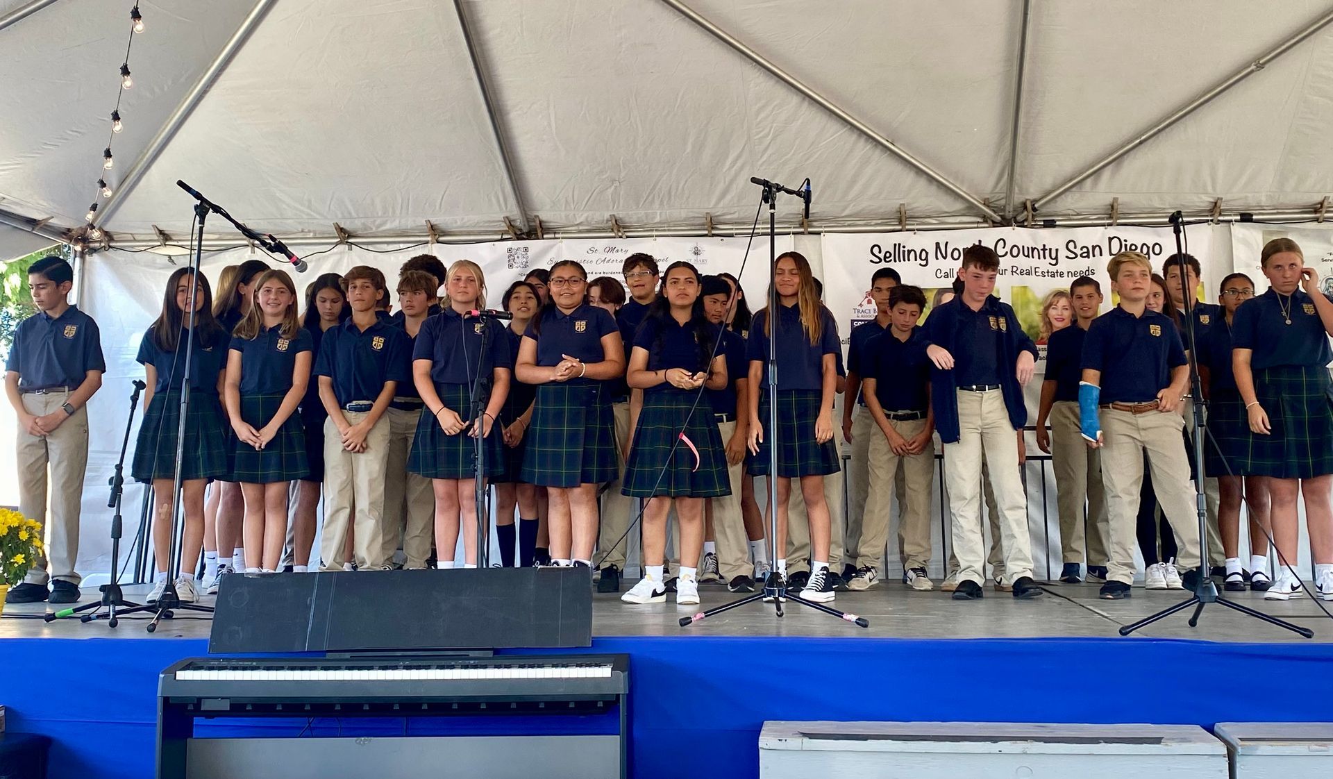 Choir group in navy and tan outfits standing on a stage under a white tent, with microphones and music stands