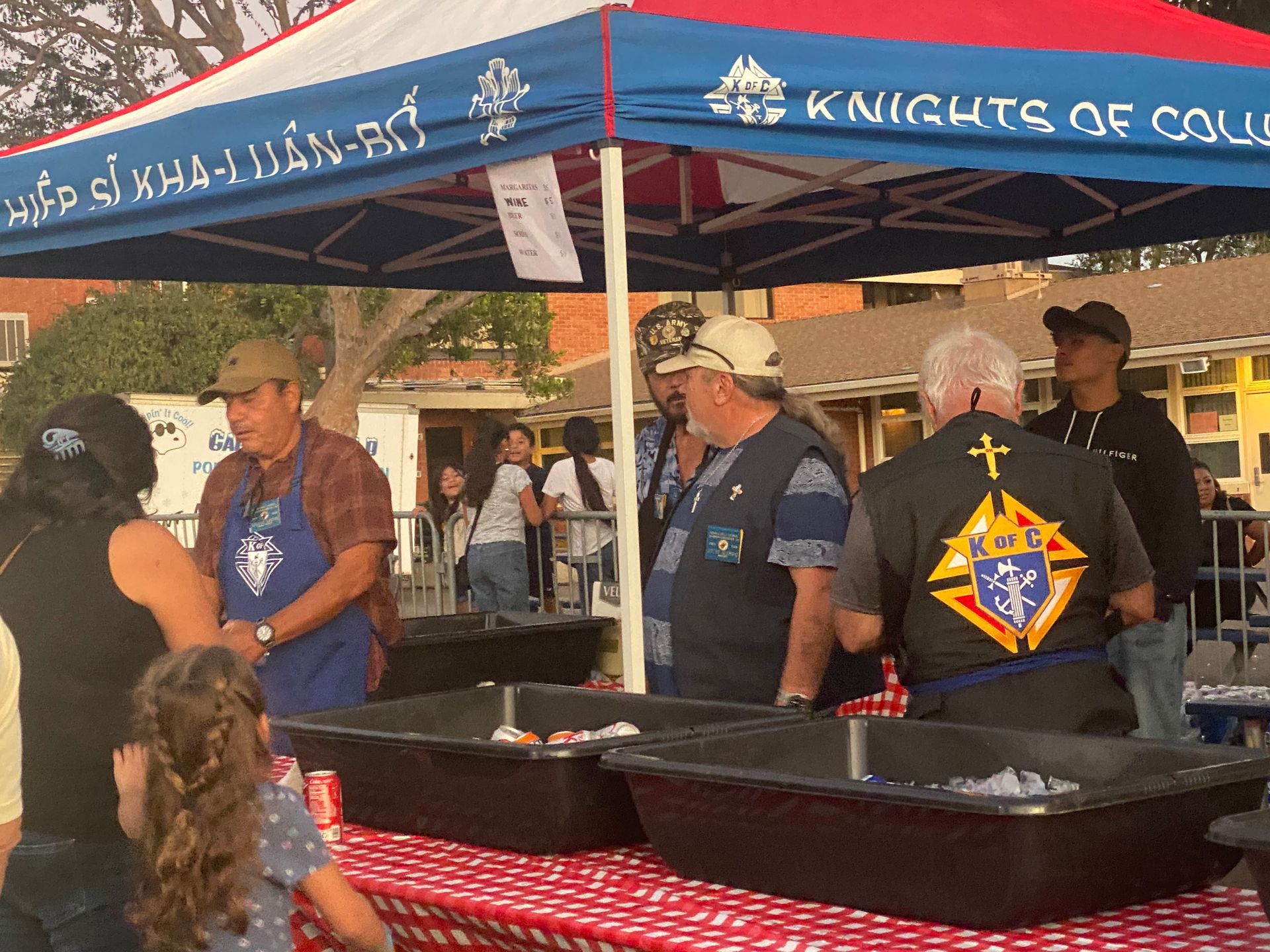 Volunteers at a Knights of Columbus food booth under a blue canopy, serving from large black pans.