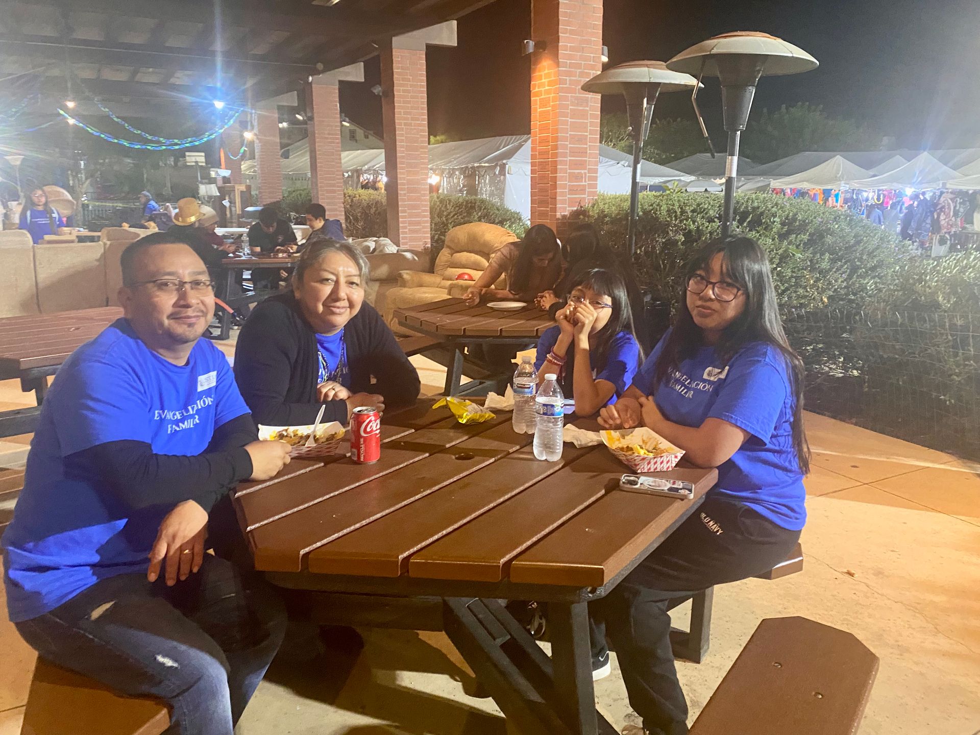 Four people in blue shirts sit at a picnic table outdoors at night, smiling and eating together.