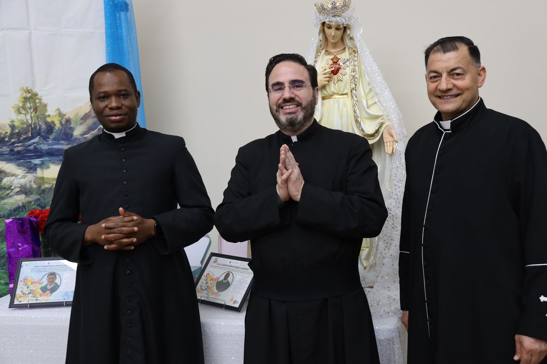 Three men in black robes pose indoors beside a statue and framed items.