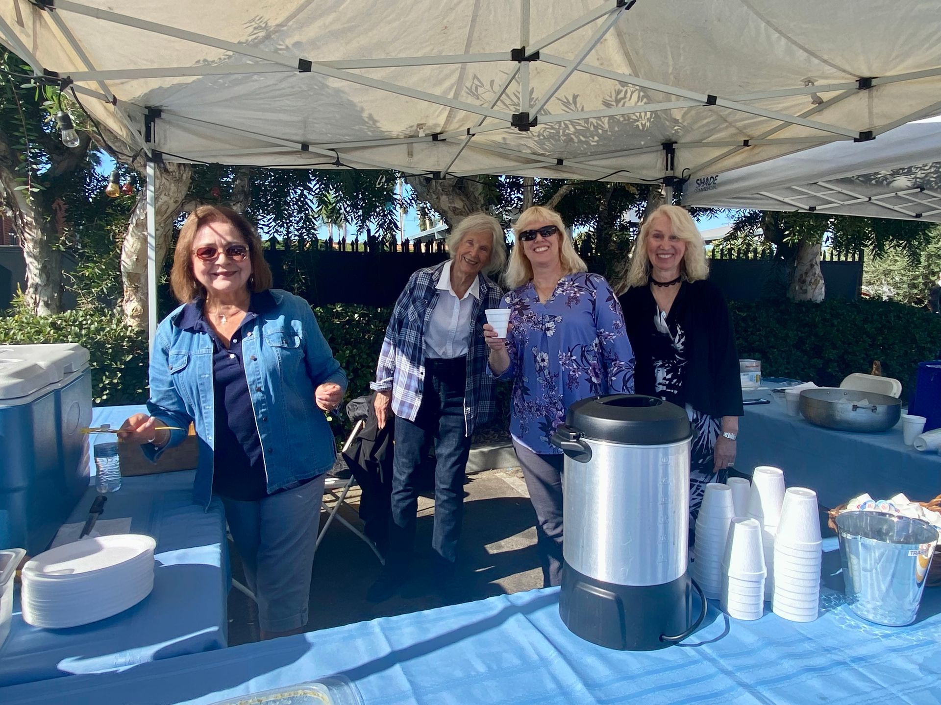 Four women standing behind an outdoor refreshment table under a white canopy.