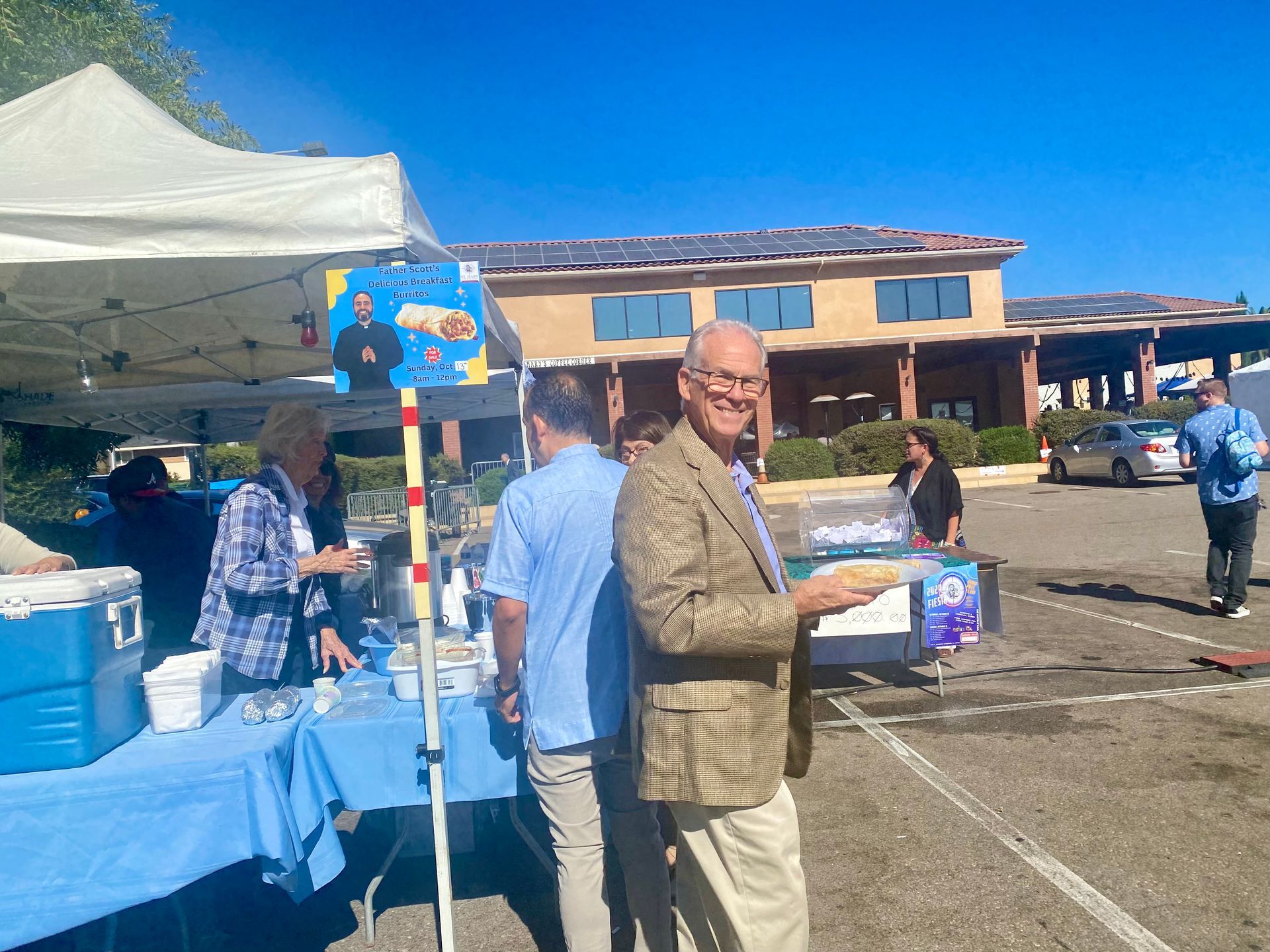 Outdoor community event with a man holding a cup near a blue table and white tent under a sunny sky