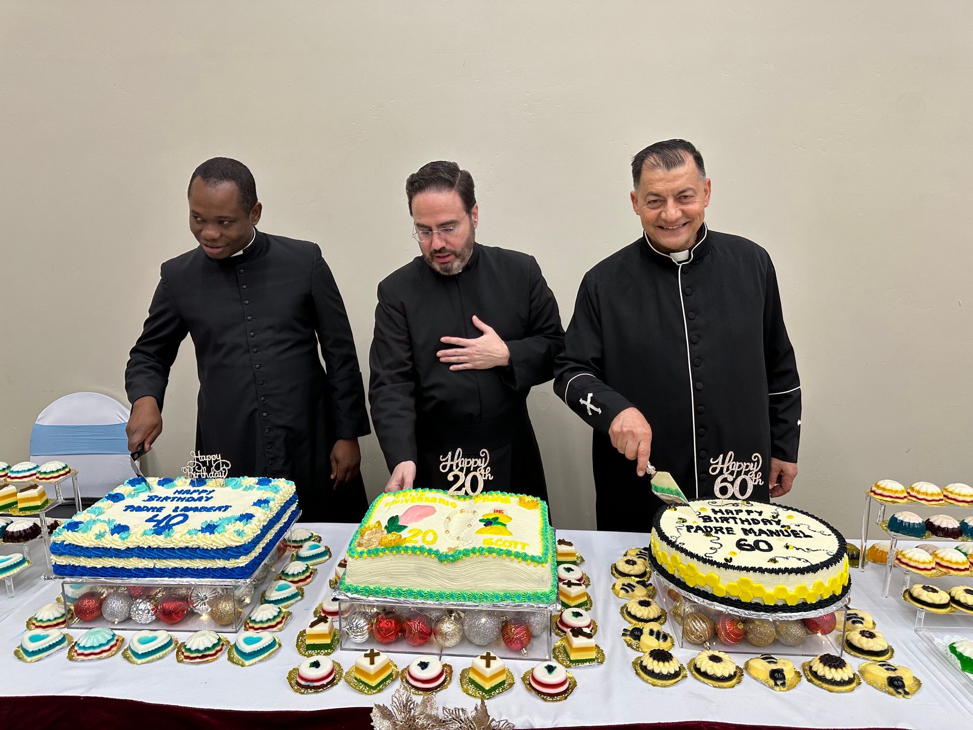 Three people stand behind a table covered with decorated cakes and cupcakes, posing at a celebratory event.