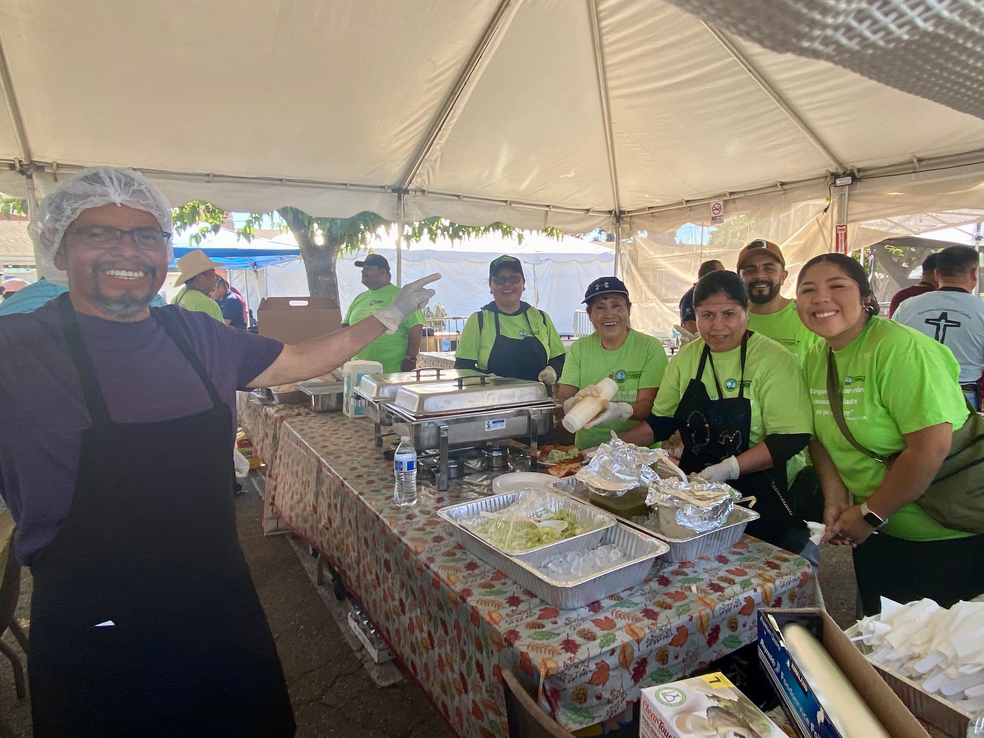 People serving food at a buffet table under a tent, with trays and chafing dishes on display.