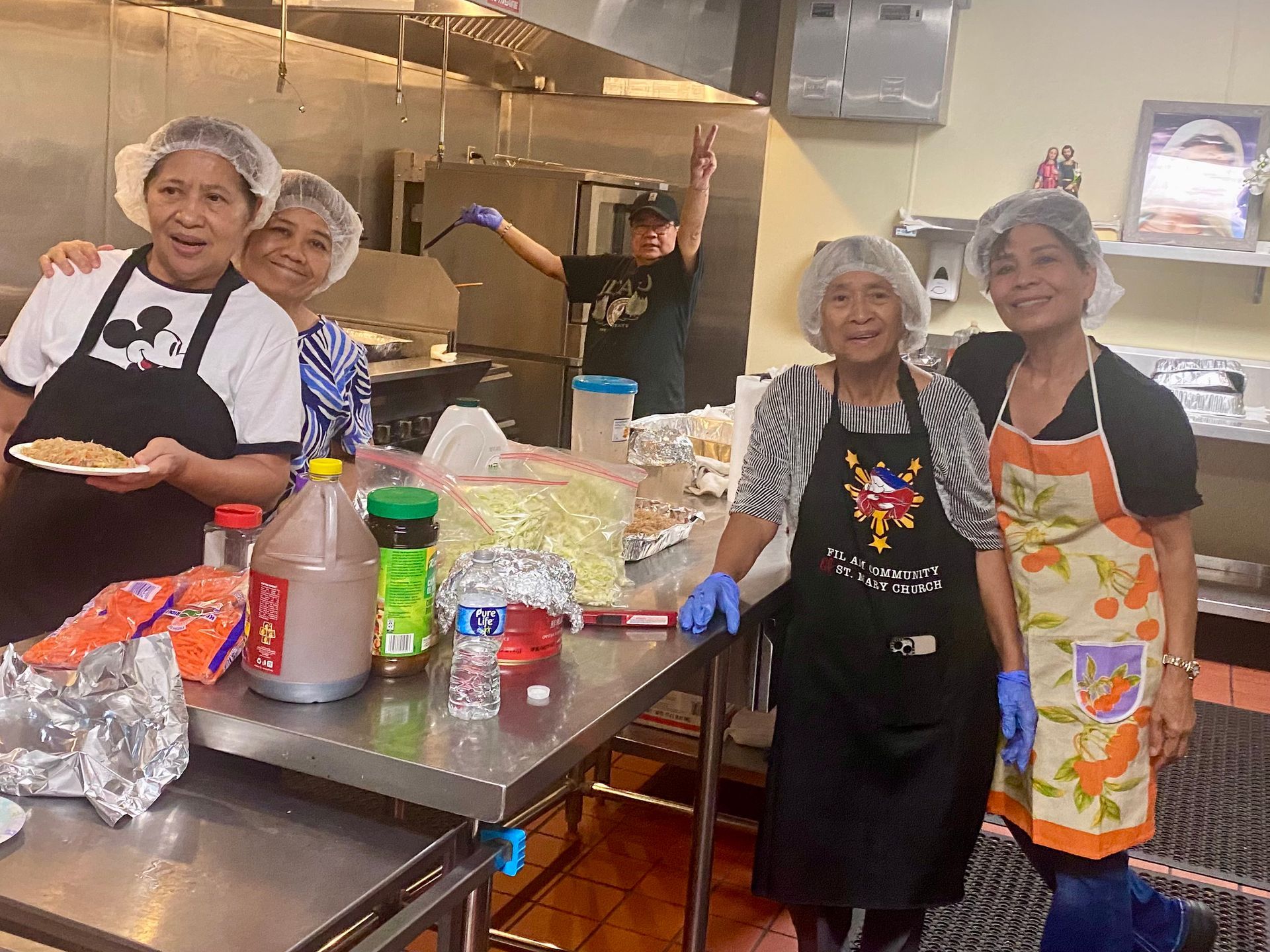 Volunteers in aprons smiling at a prep table in a kitchen, with ingredients and utensils spread out