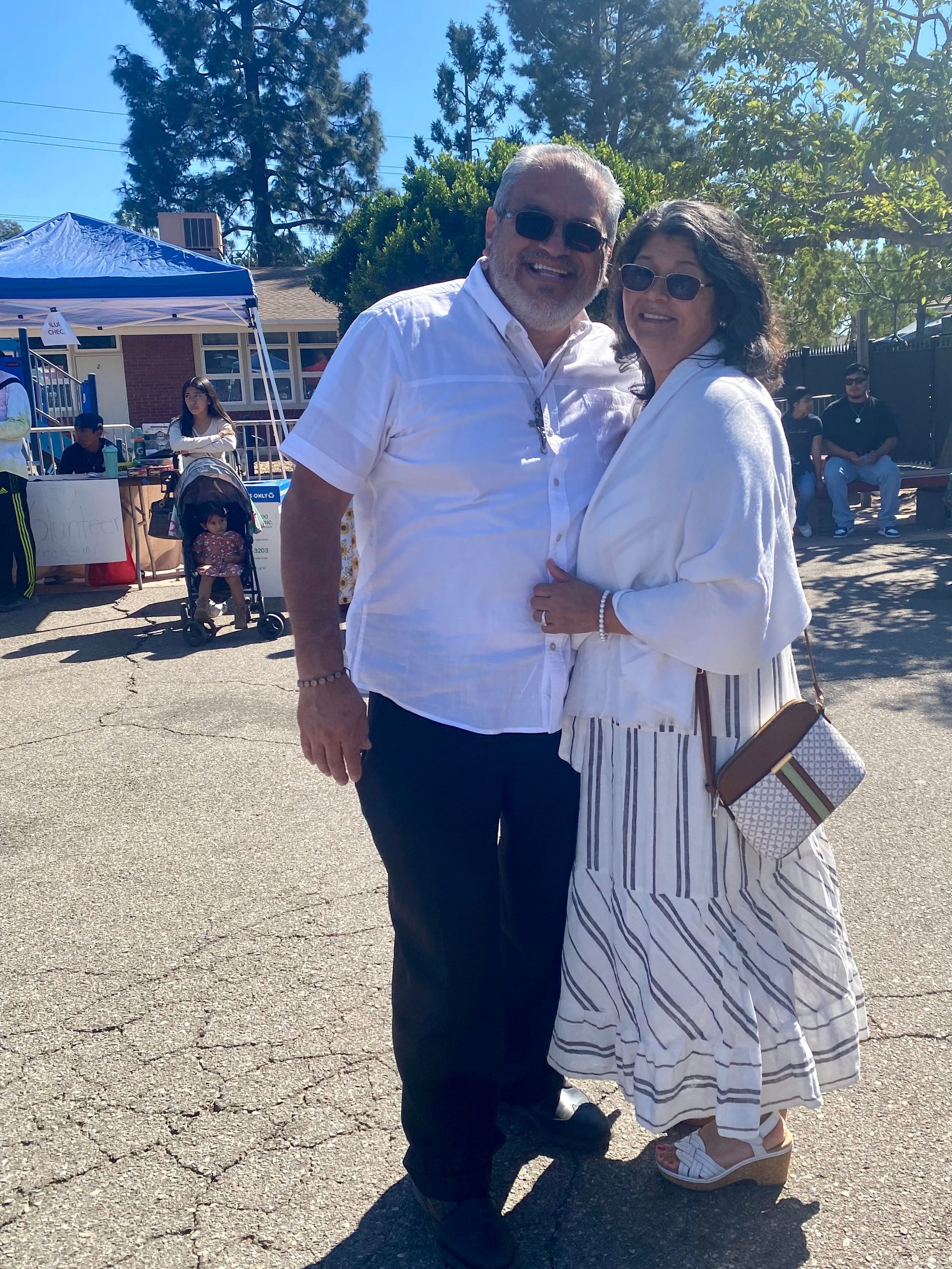 Two people smiling outdoors at a sunny market, standing close together in white clothing