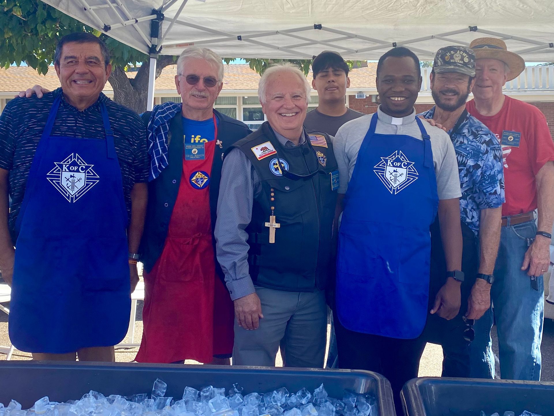Group of people posing under a canopy, several in blue aprons, at an outdoor event.