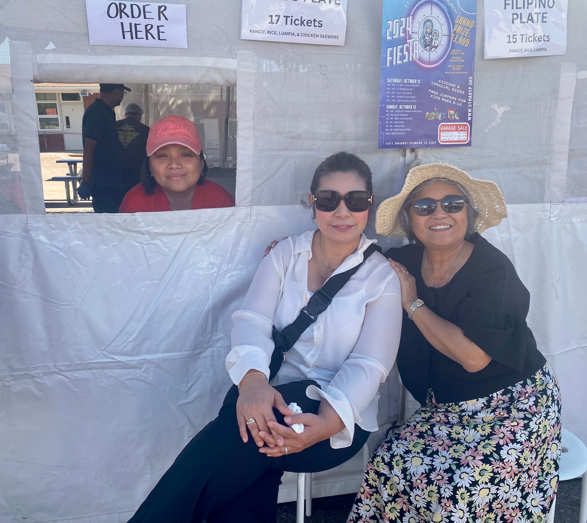 Three people smiling at a festival food booth, with menu signs and a vendor in the background.