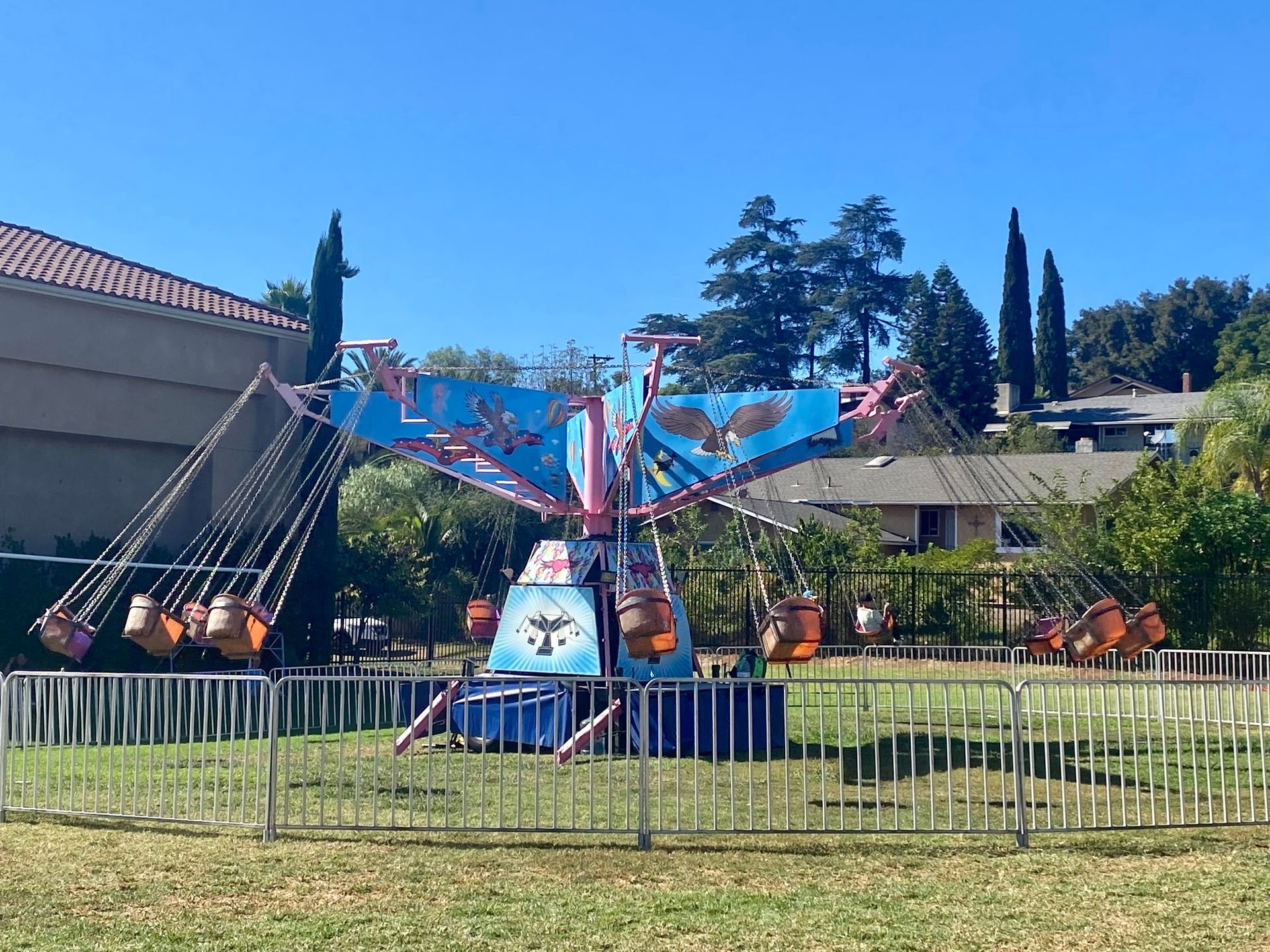 Small amusement park ride with colorful seats and a white fence, set against houses and trees under a blue sky