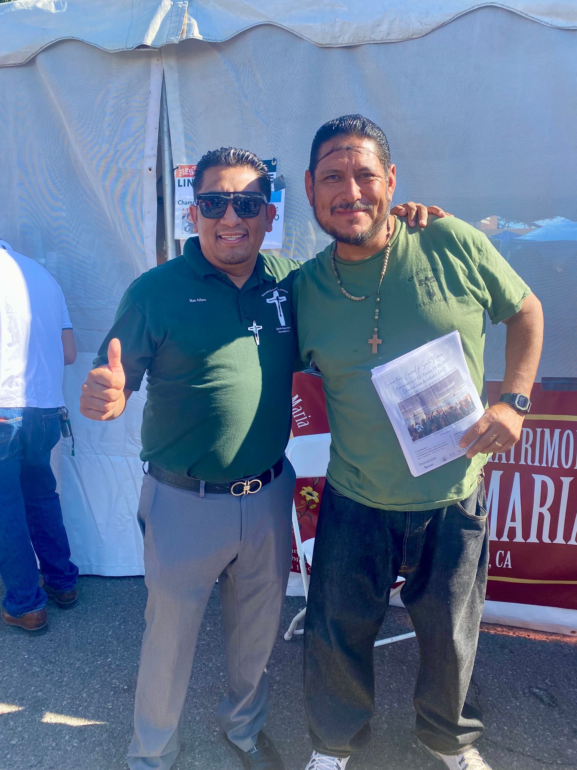 Two men posing outdoors at an event, smiling with arms around each other, holding a flyer.