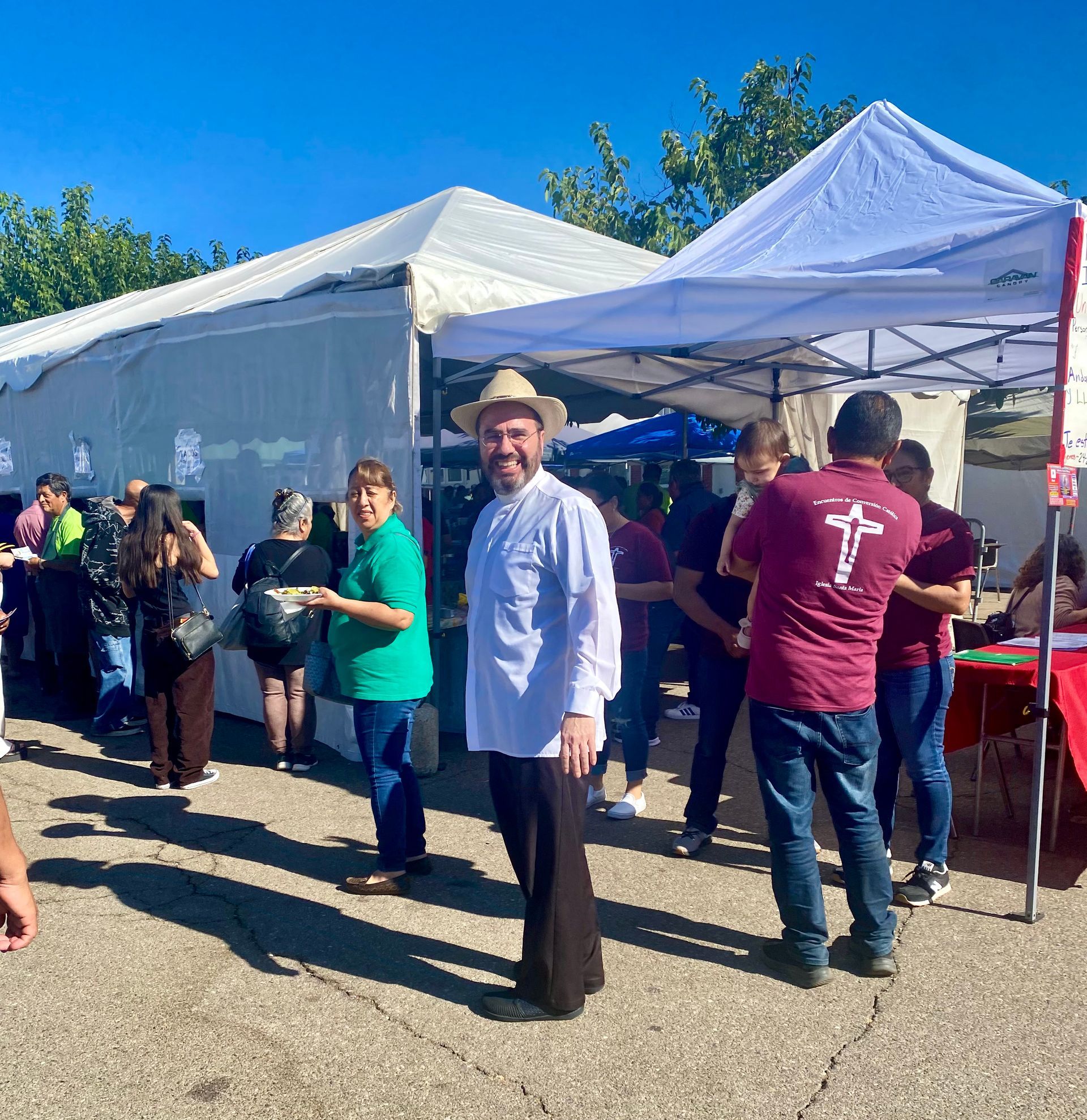 People gathered under white tents at an outdoor market, with a man in a white shirt smiling in the foreground.