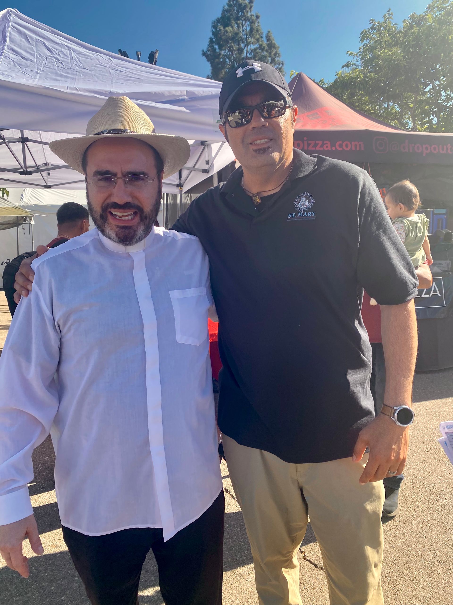 Two men standing outdoors at a market, one in a white shirt and straw hat, the other in a black polo and cap.
