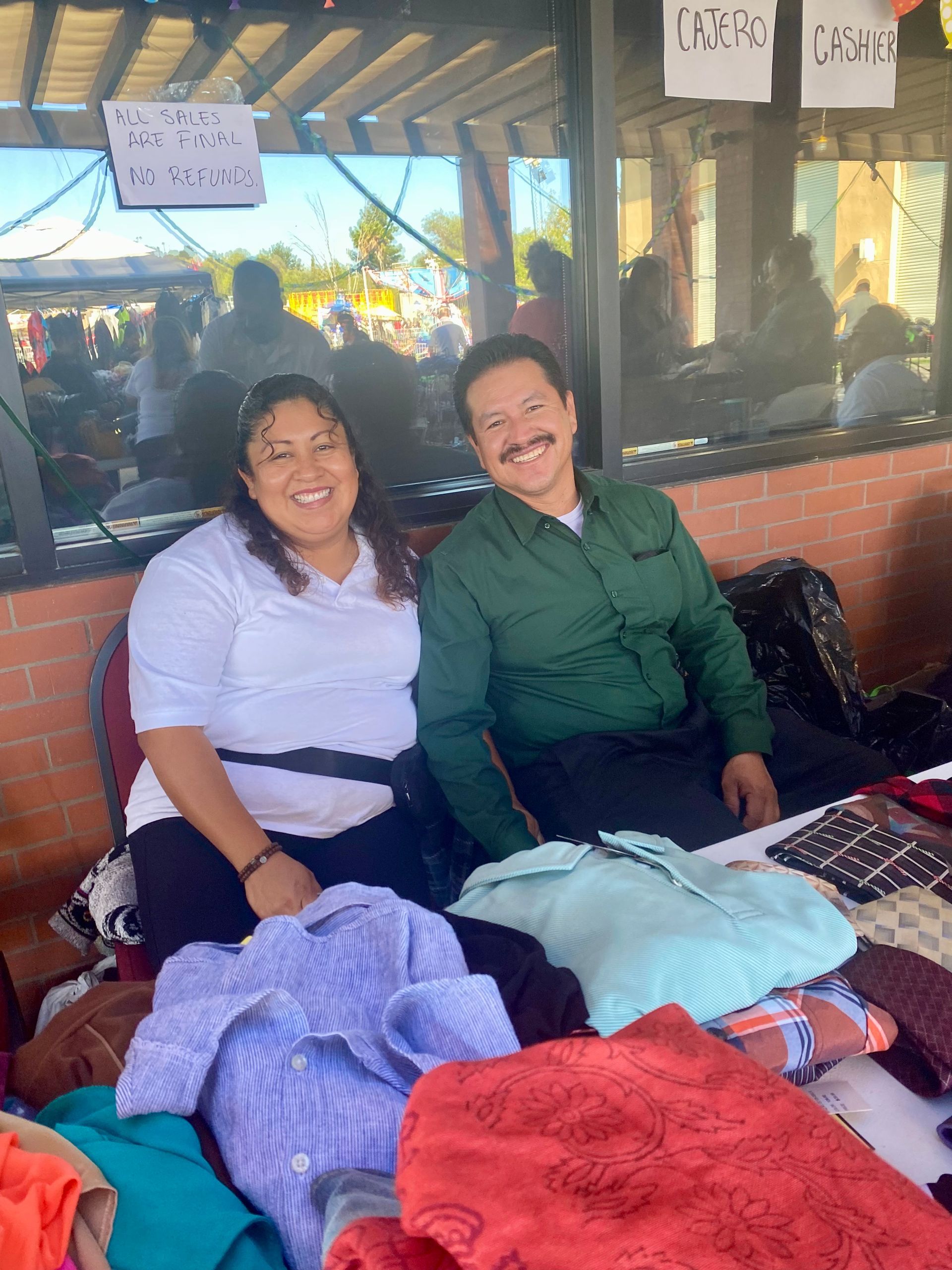 Two people sitting by a table of folded clothes, smiling in a market stall.