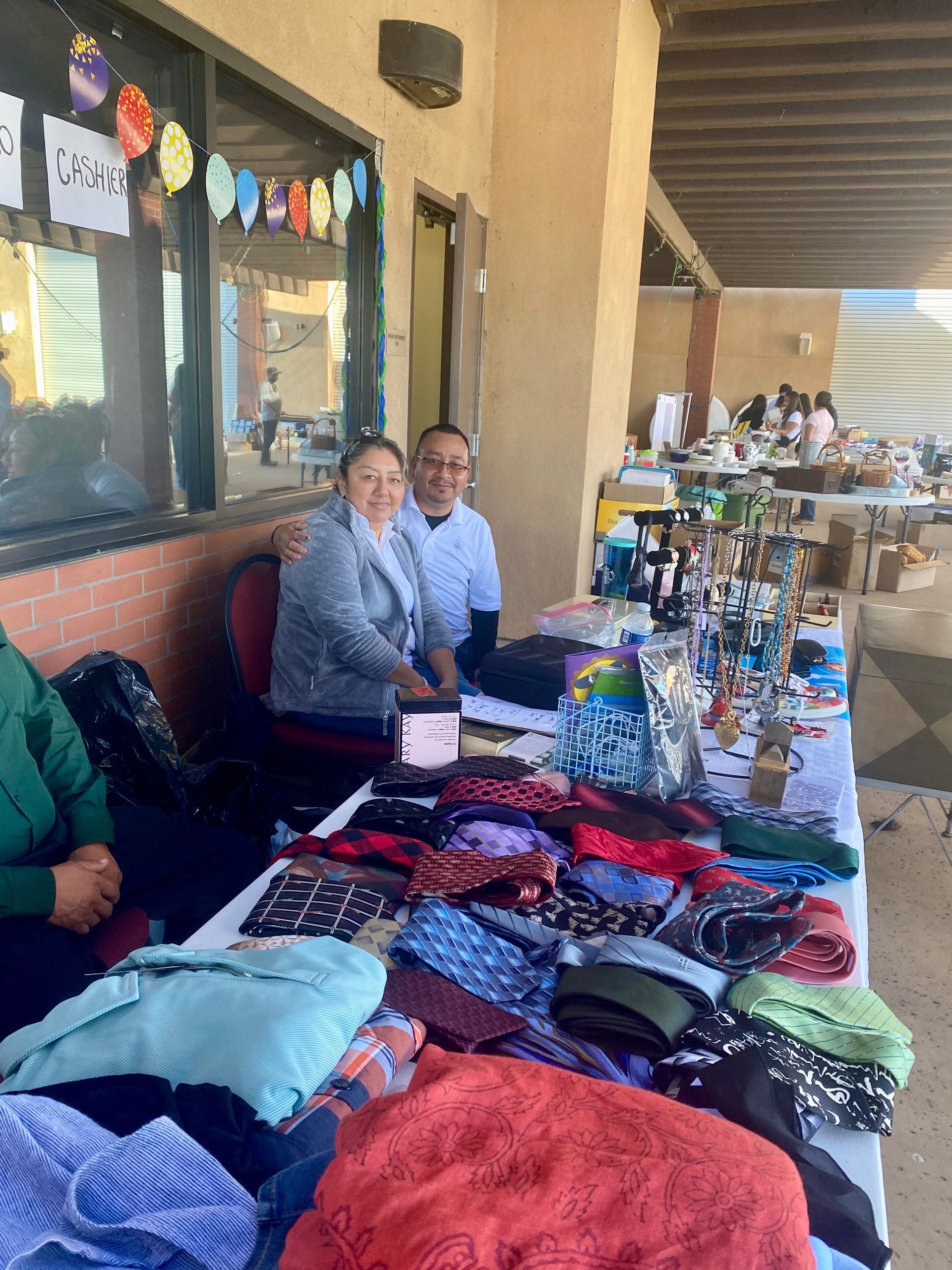 Two people behind a market table displaying folded sweaters, hats, and accessories outdoors