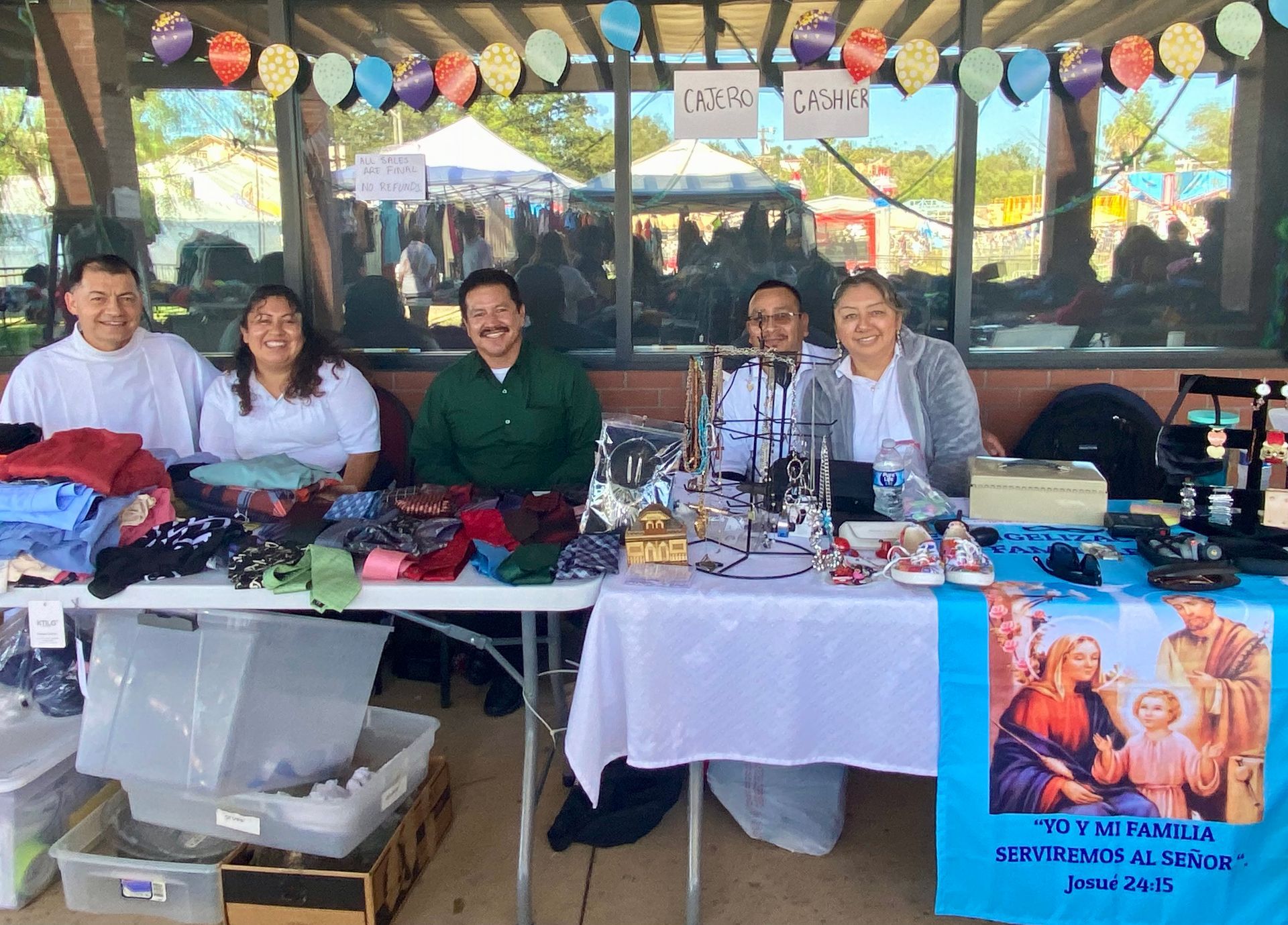 Five people sit behind a vendor table with crafts and decorations under a festive canopy.