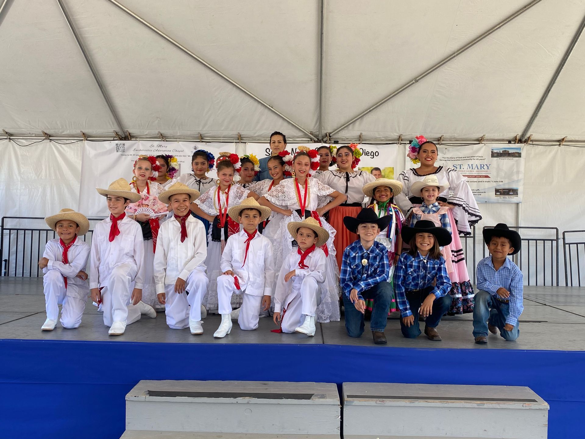 Children in colorful costumes pose on a stage under a white tent, arranged in two rows.