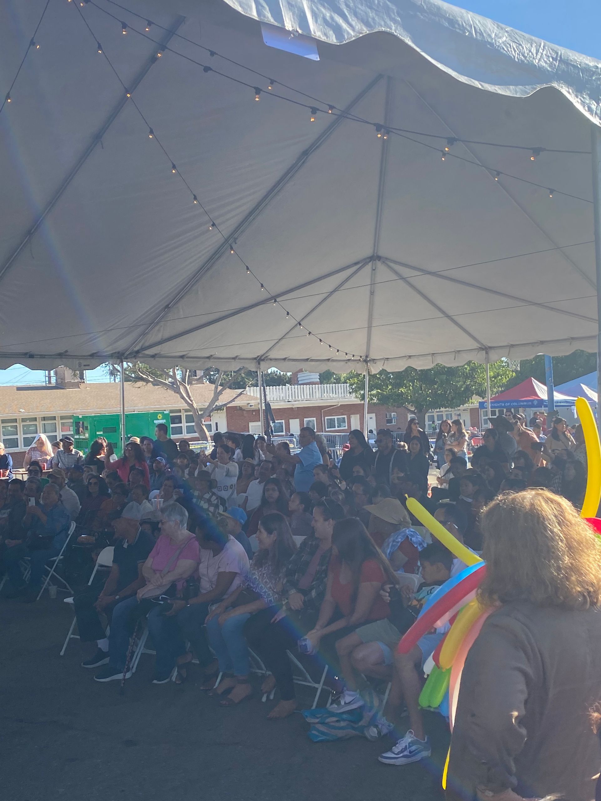 Crowd seated under a large white canopy at an outdoor event, with colorful streamers in the foreground.
