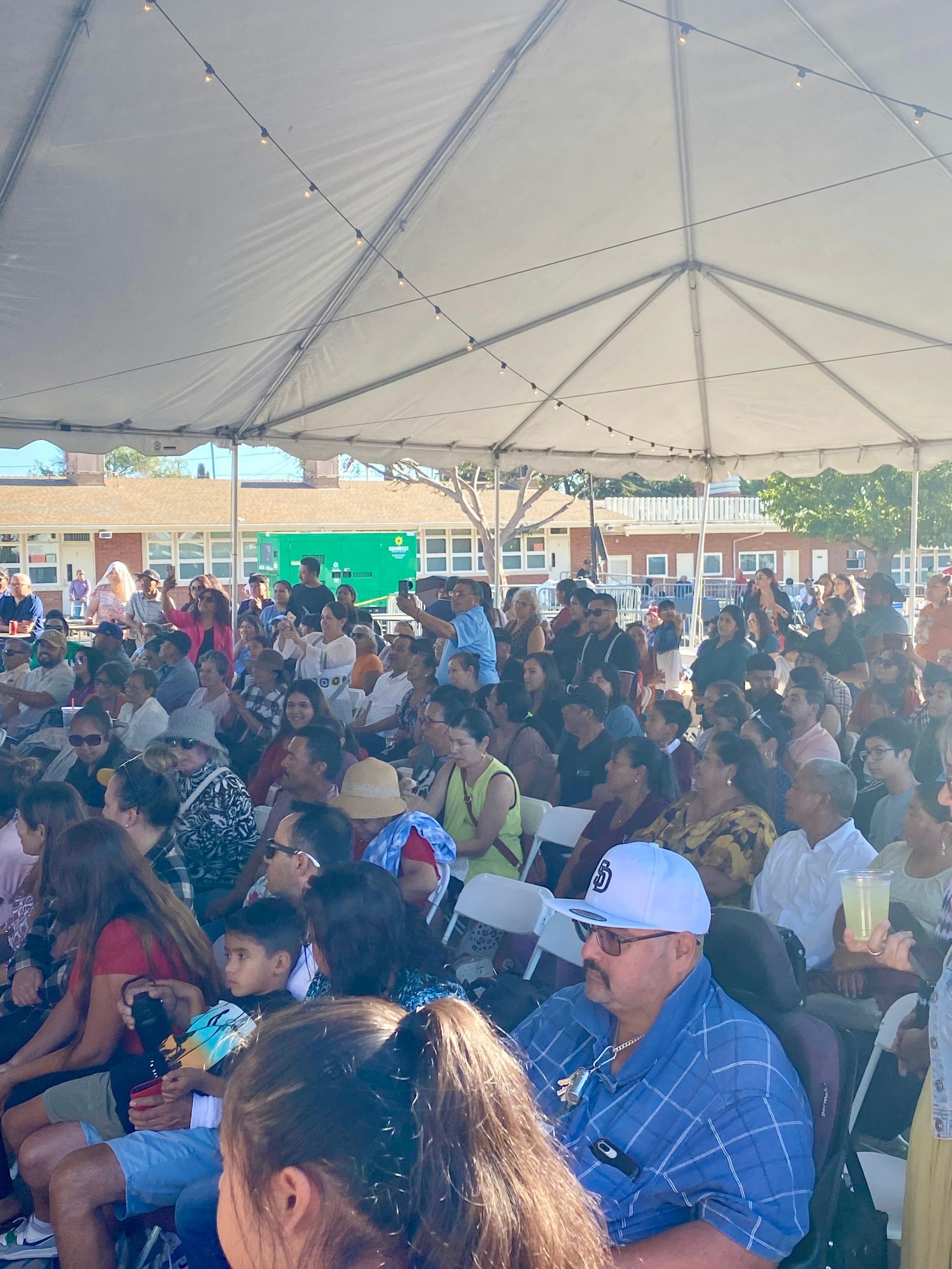 Crowd seated under a white tent at an outdoor event, with people facing a stage.