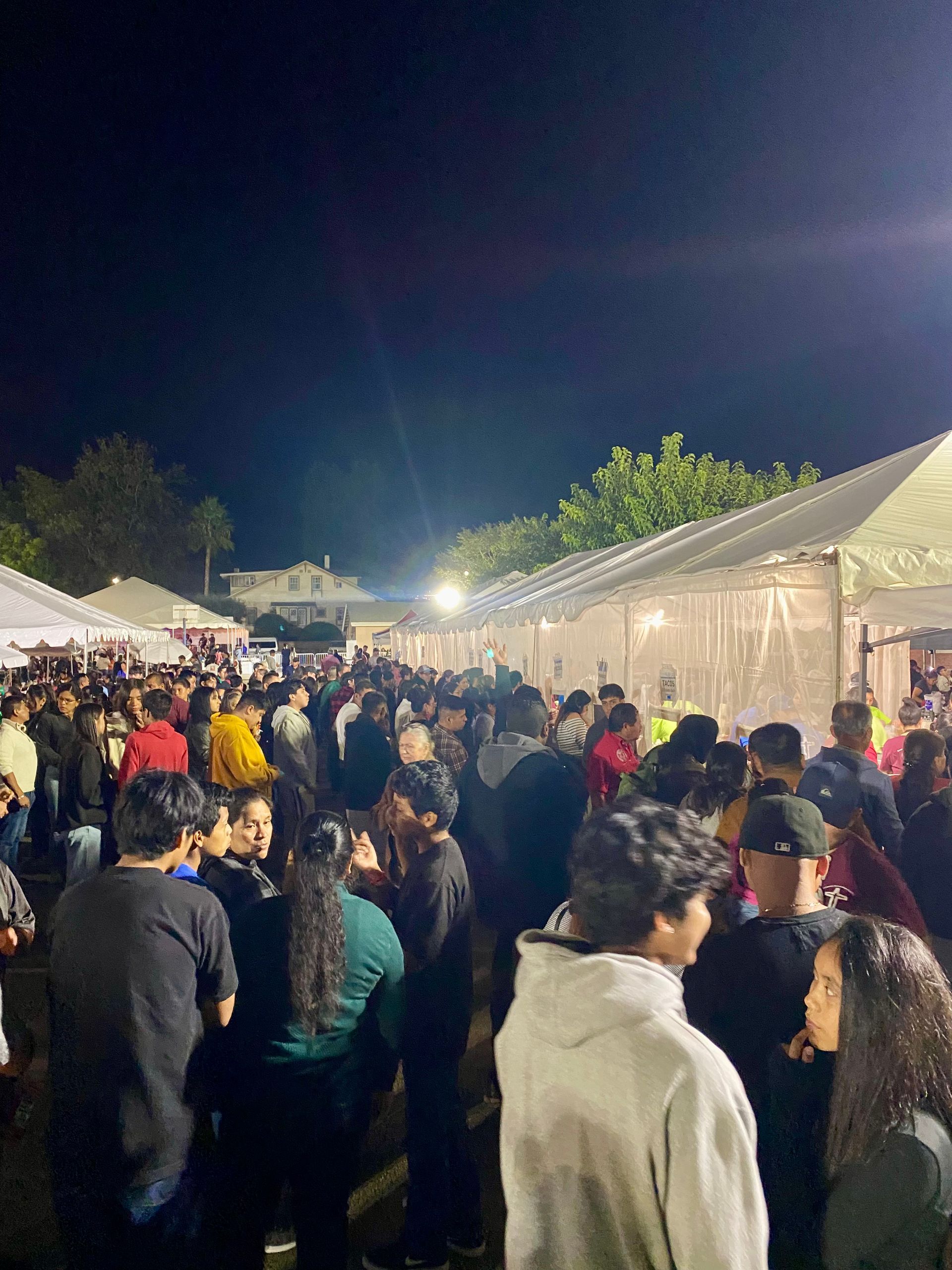Crowded outdoor night market beside white tents, with people walking under bright lights.