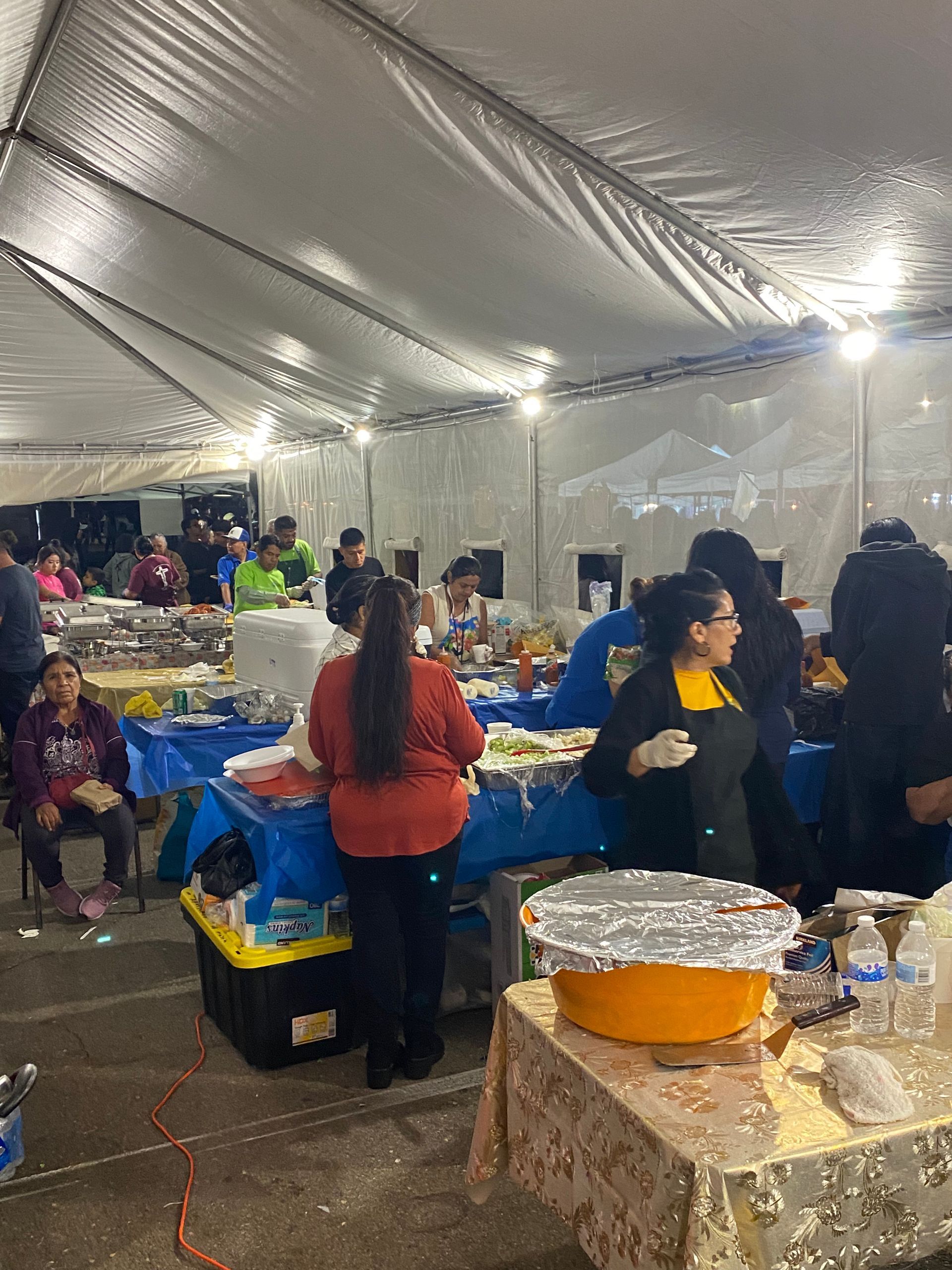 People serving food at a buffet under a large white tent with blue table coverings.