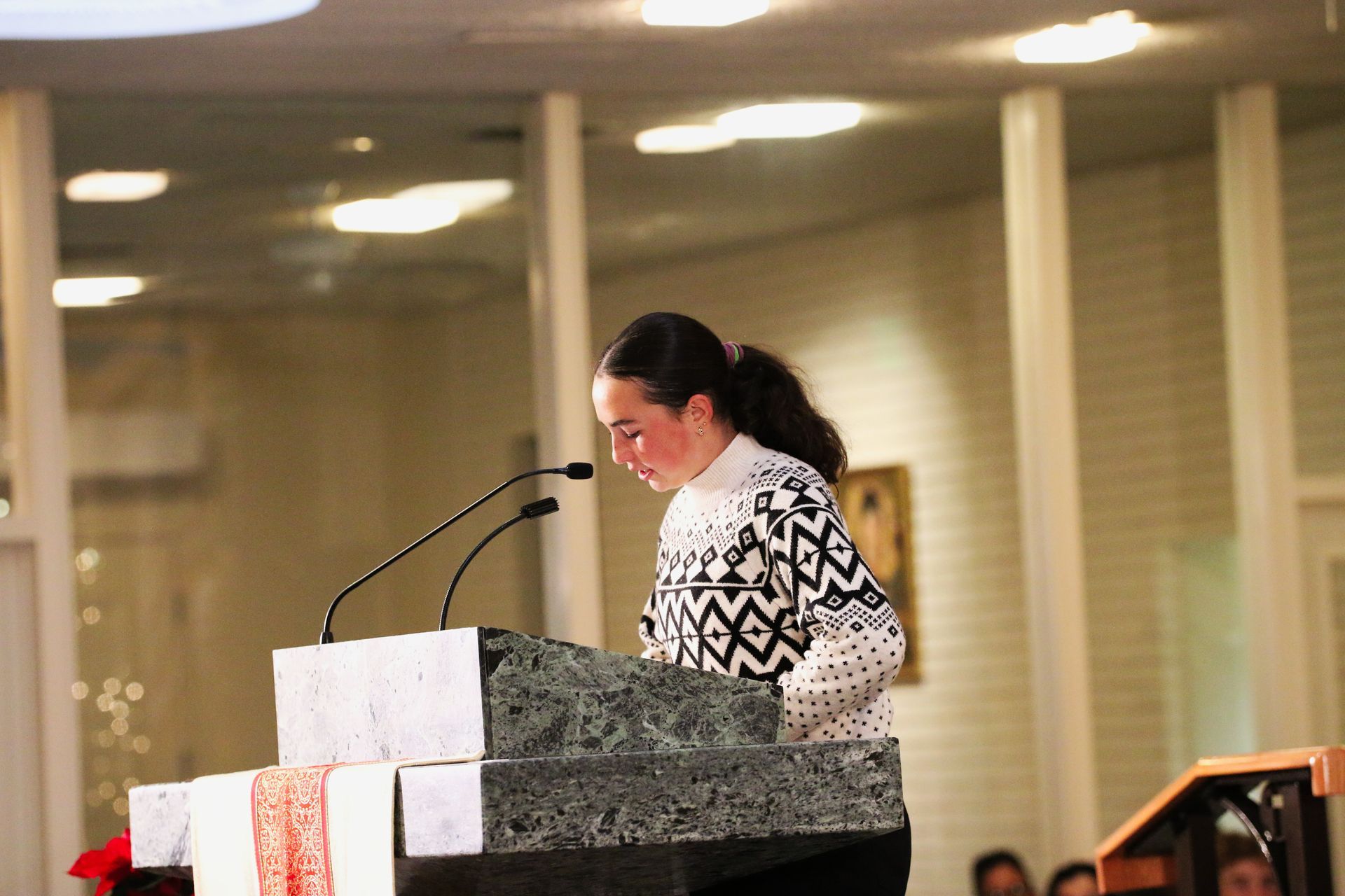 Woman speaking at a podium with microphones in a warmly lit indoor hall