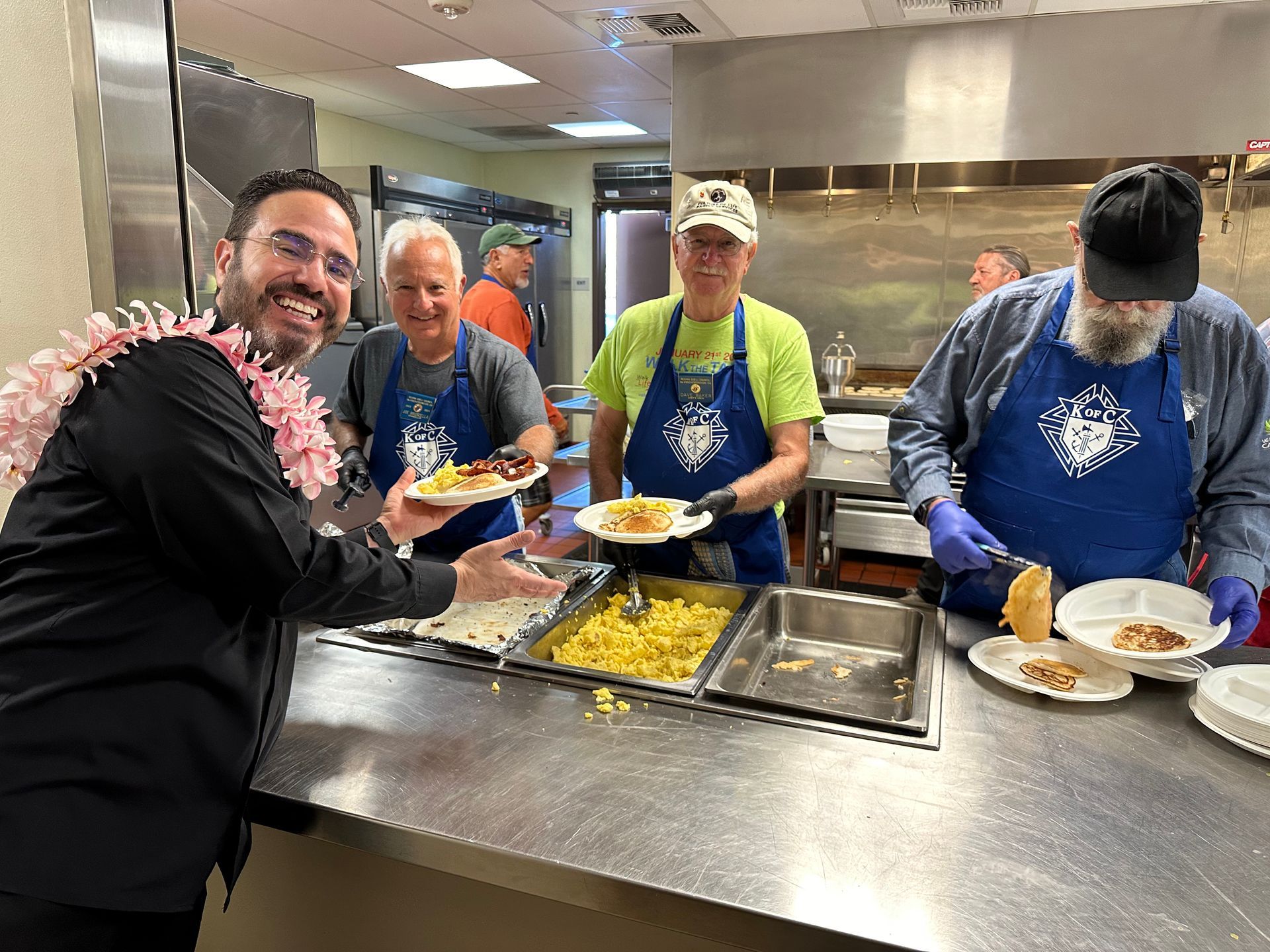 Volunteers serving food at a stainless-steel cafeteria counter, with trays of pasta and plates ready