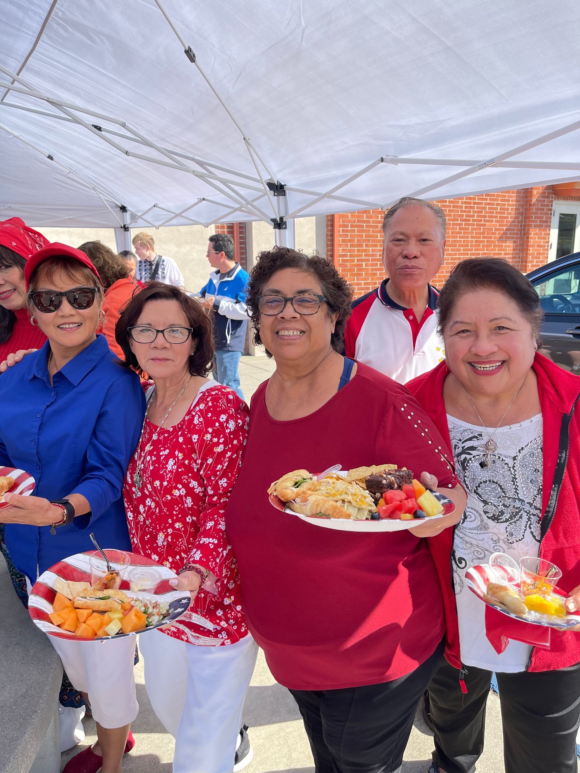 Smiling group under a canopy, holding plates of food at an outdoor gathering.