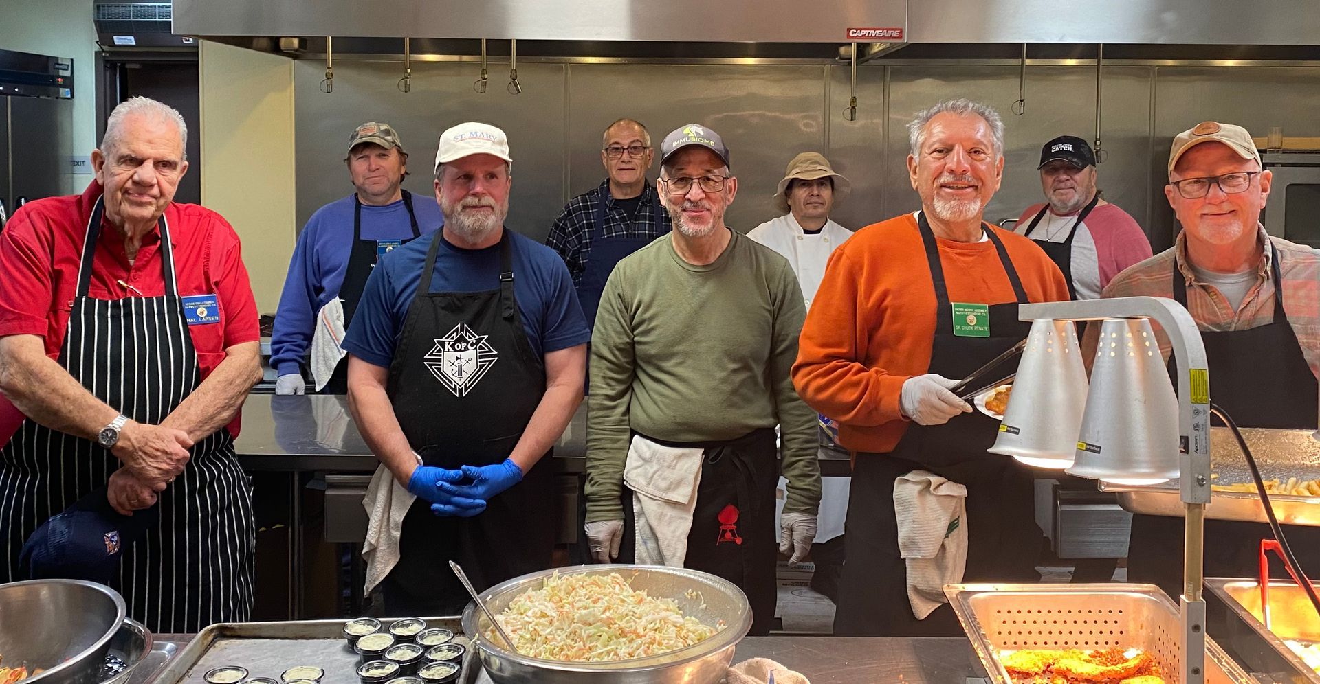 Five people in aprons and gloves stand behind a buffet line in a kitchen, smiling at the camera.