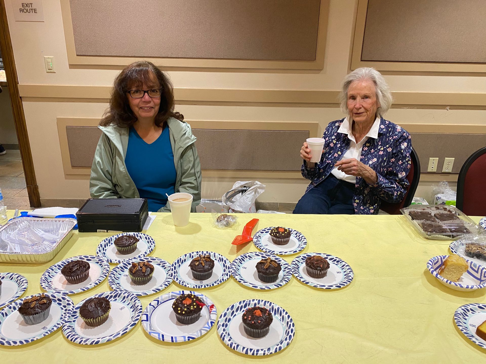 Two women at a table with plates of cupcakes in a banquet room, one holding a cupcake.