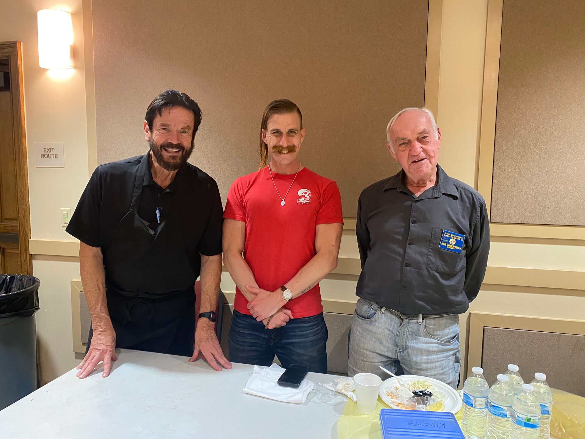 Three people standing behind a table with papers and water bottles at an indoor event table