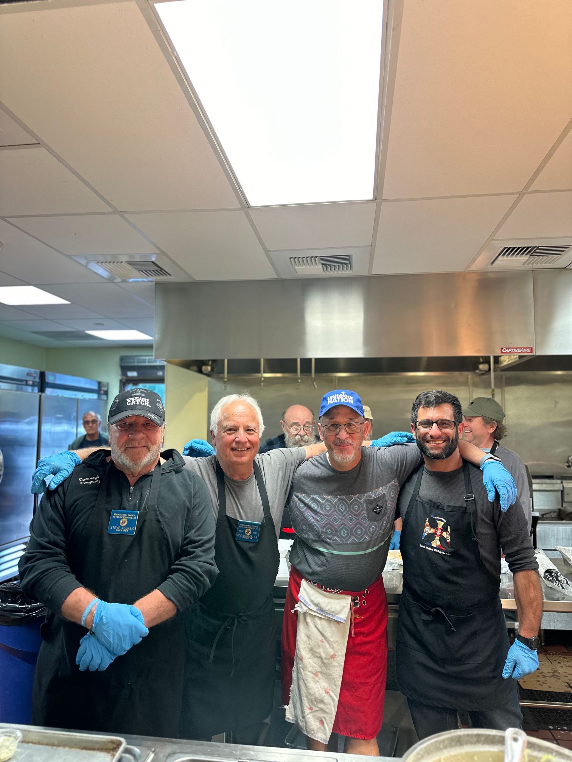 Five people in chef uniforms and gloves stand together in a kitchen, smiling at the camera.