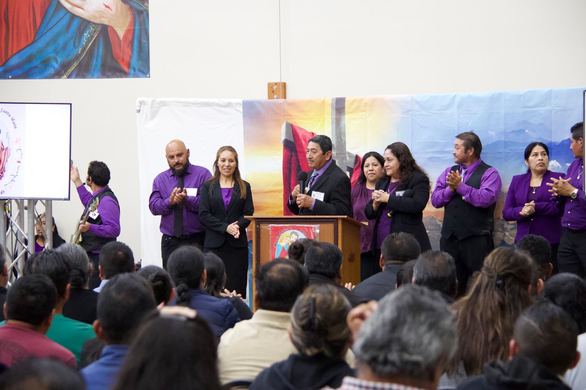 People on a stage speaking at a podium before a seated audience, with purple-clad participants nearby