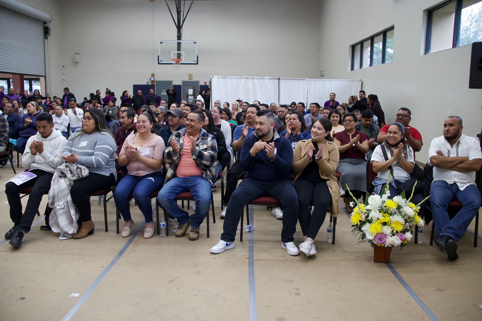Audience seated in a gymnasium, facing forward, with flowers at the front right and a basketball hoop above.