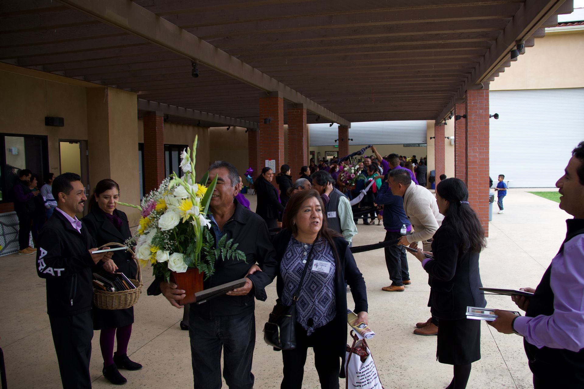 People carrying floral wreaths and standing in a covered outdoor corridor during a procession