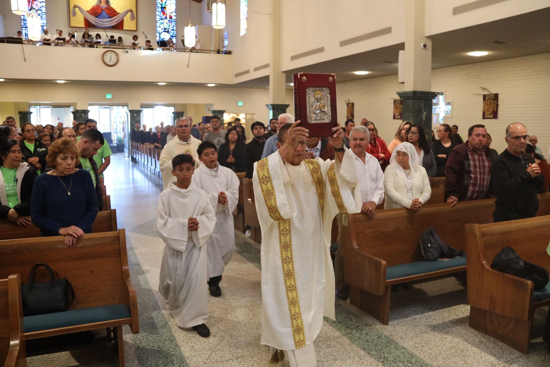 Priest in white and gold leads a church procession down the aisle as congregants stand in the pews.