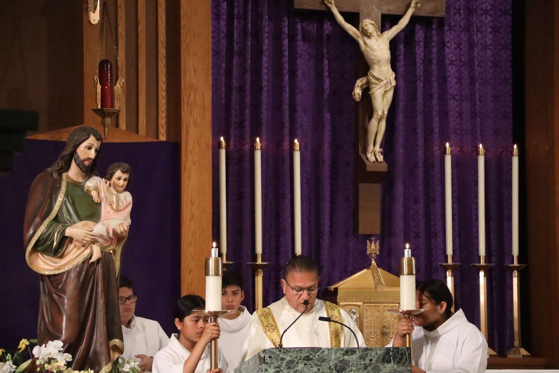 Priest and altar servers at a church altar beneath a crucifix and candles, with a statue of Mary and child.