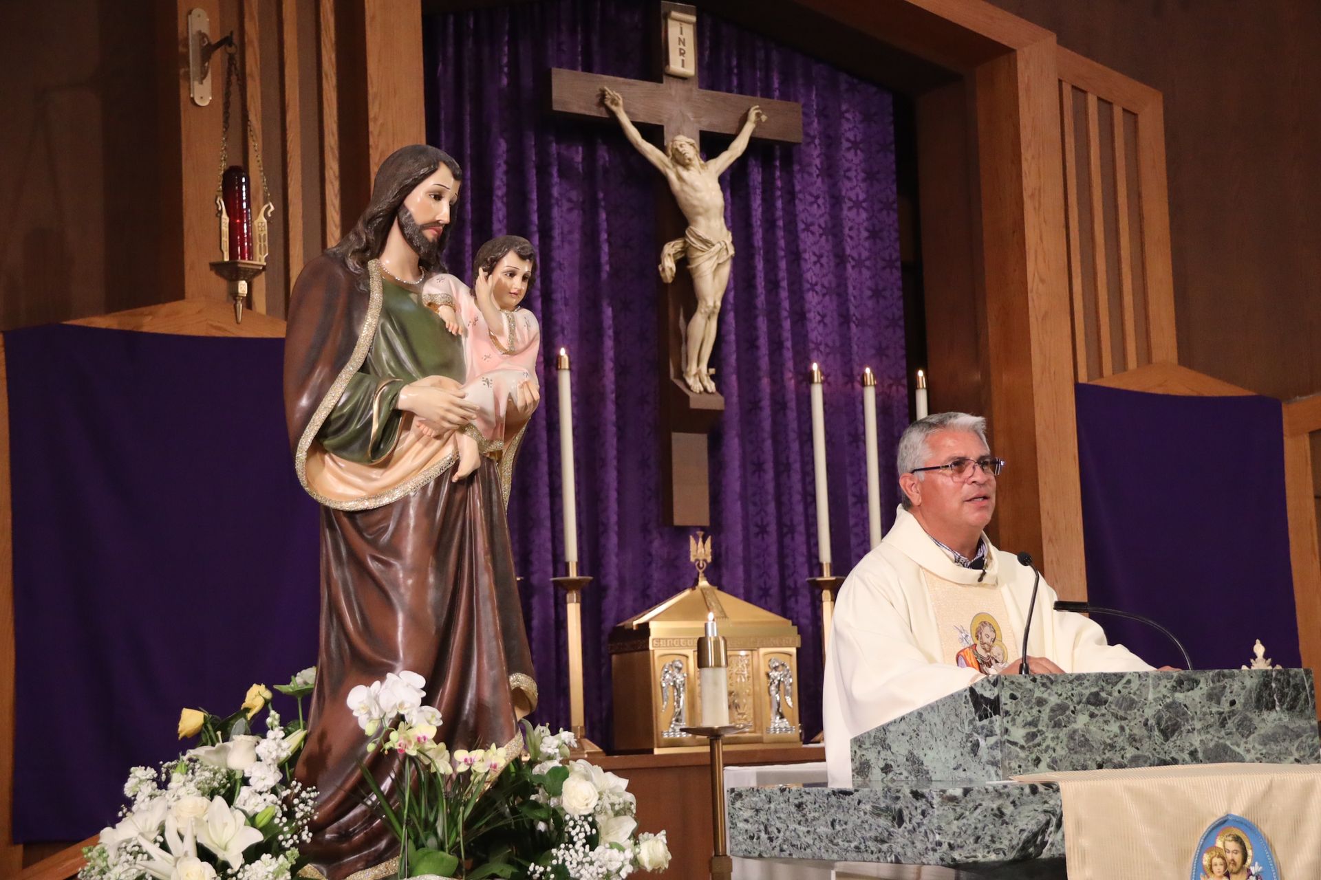Church altar with priest at lectern, crucifix, and statue of Mary holding child, framed by purple drapes