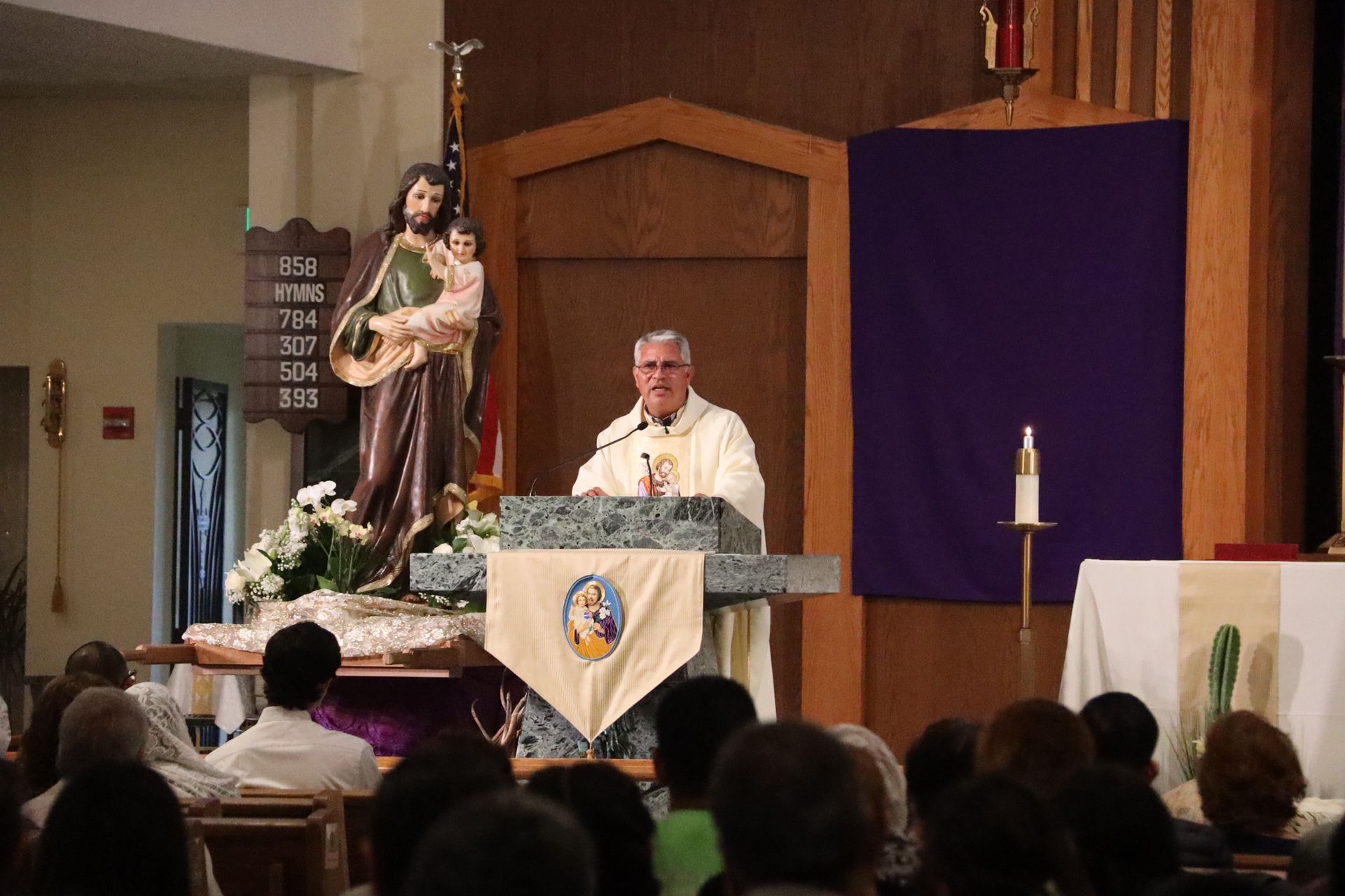 Priest speaking at church altar beside statue of Mary and purple Lenten drape