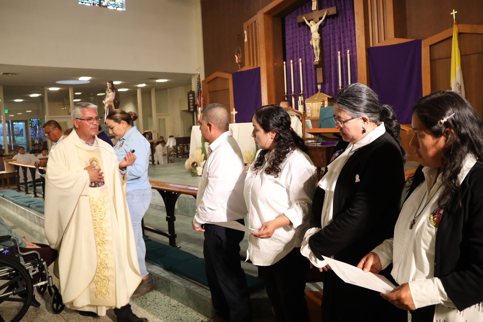 Priest in white vestments greets a line of people inside a church by the altar