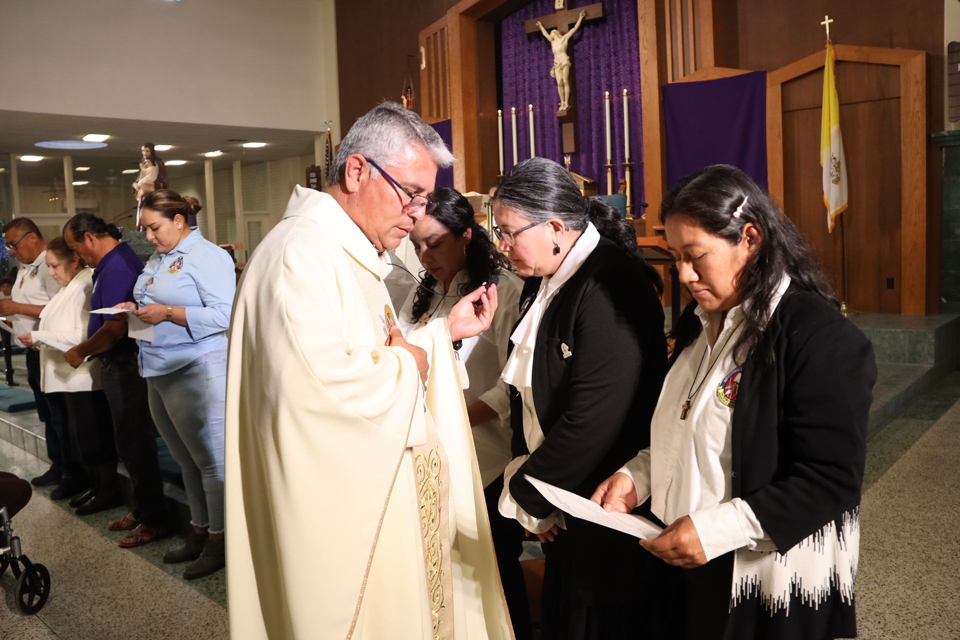 Priest blessing two people in a church, with a crucifix and purple drapery in the background