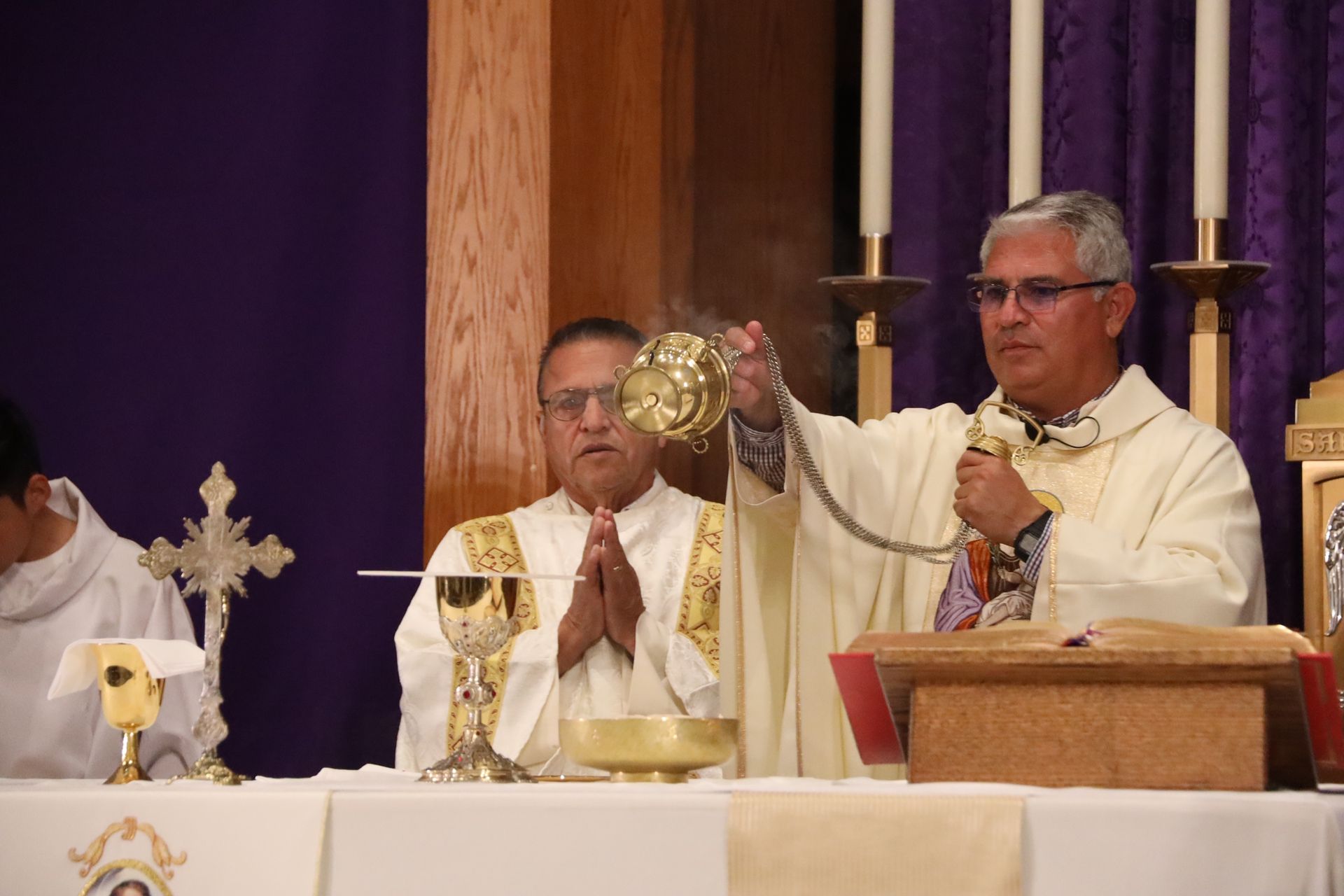 Priest elevates a golden chalice during Mass at an altar with candles and a seated clergy member
