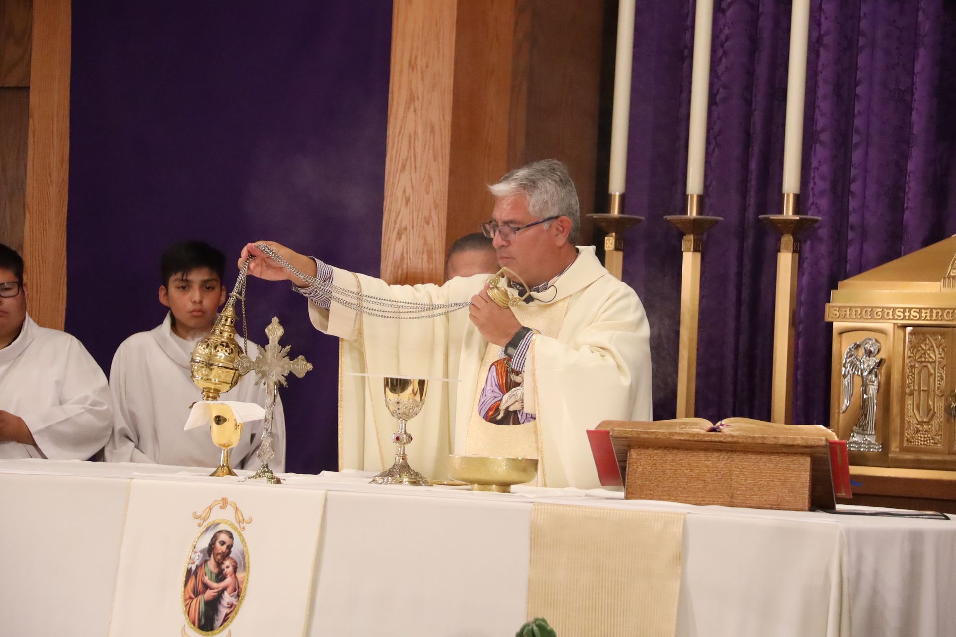 Priest elevates the Eucharist at an altar with chalice, candles, and purple drapery in a church service