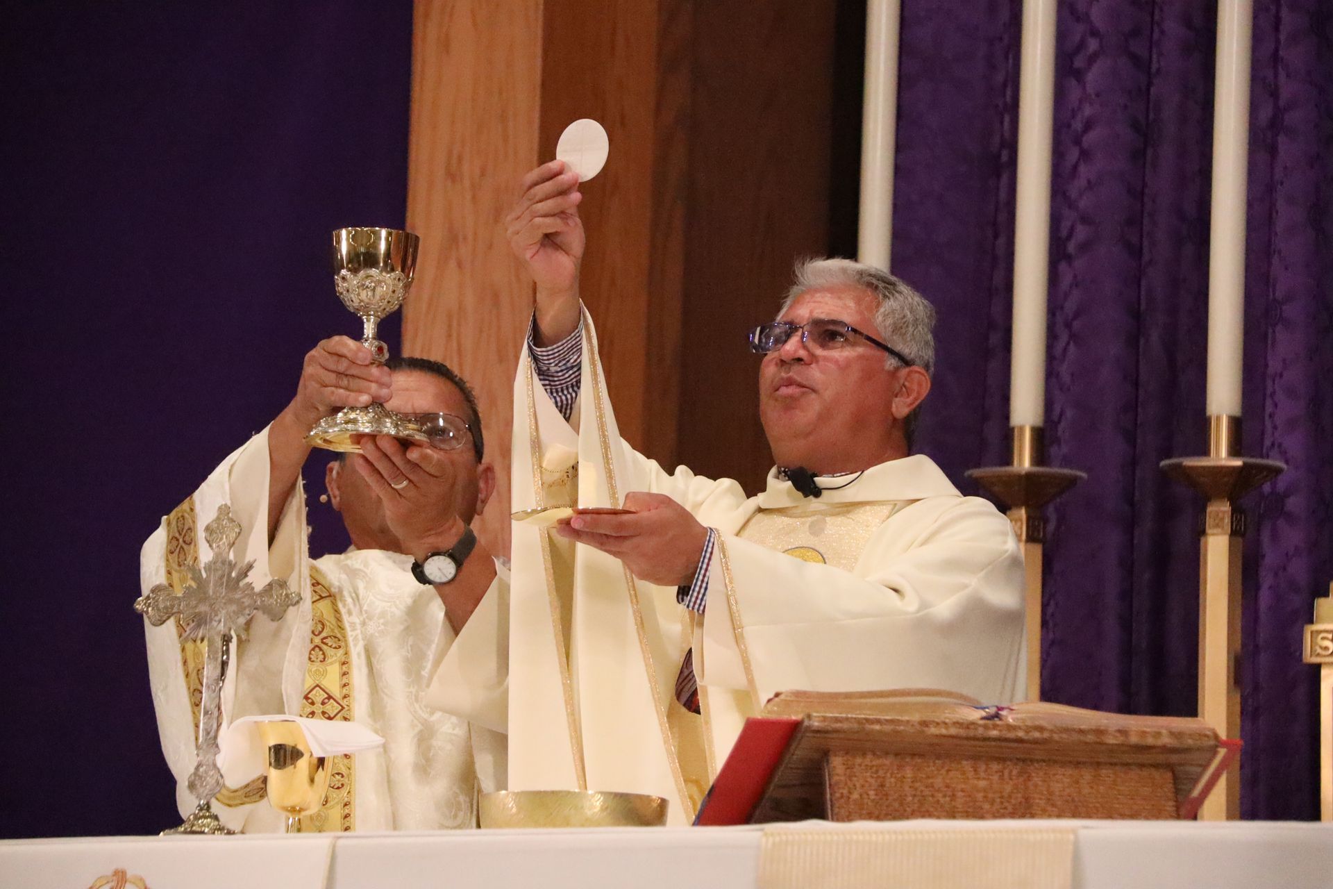 Two clergy in white robes elevating communion elements at a church altar