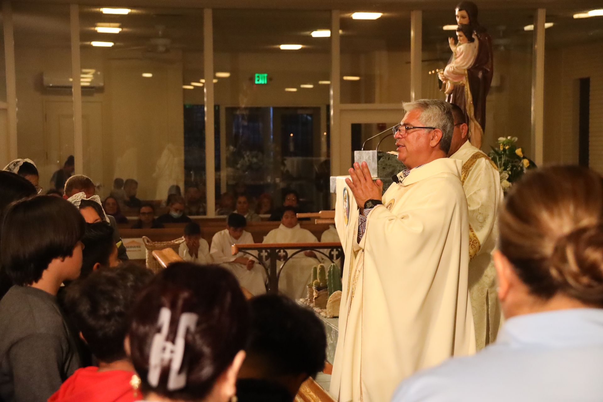 Priest in white robes speaking to a seated crowd indoors, with a statue behind him.